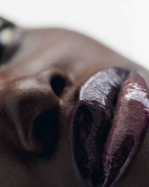 Close-up shot of an adult woman's face with dark skin and glossy purple lipstick, focusing on the lips and nose against a white background.