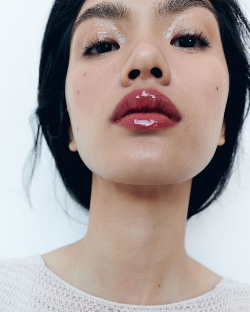 Beauty shot of a dark-haired Asian woman with glossy lips and dewy skin, gazing directly at the camera in a brightly lit indoor space.