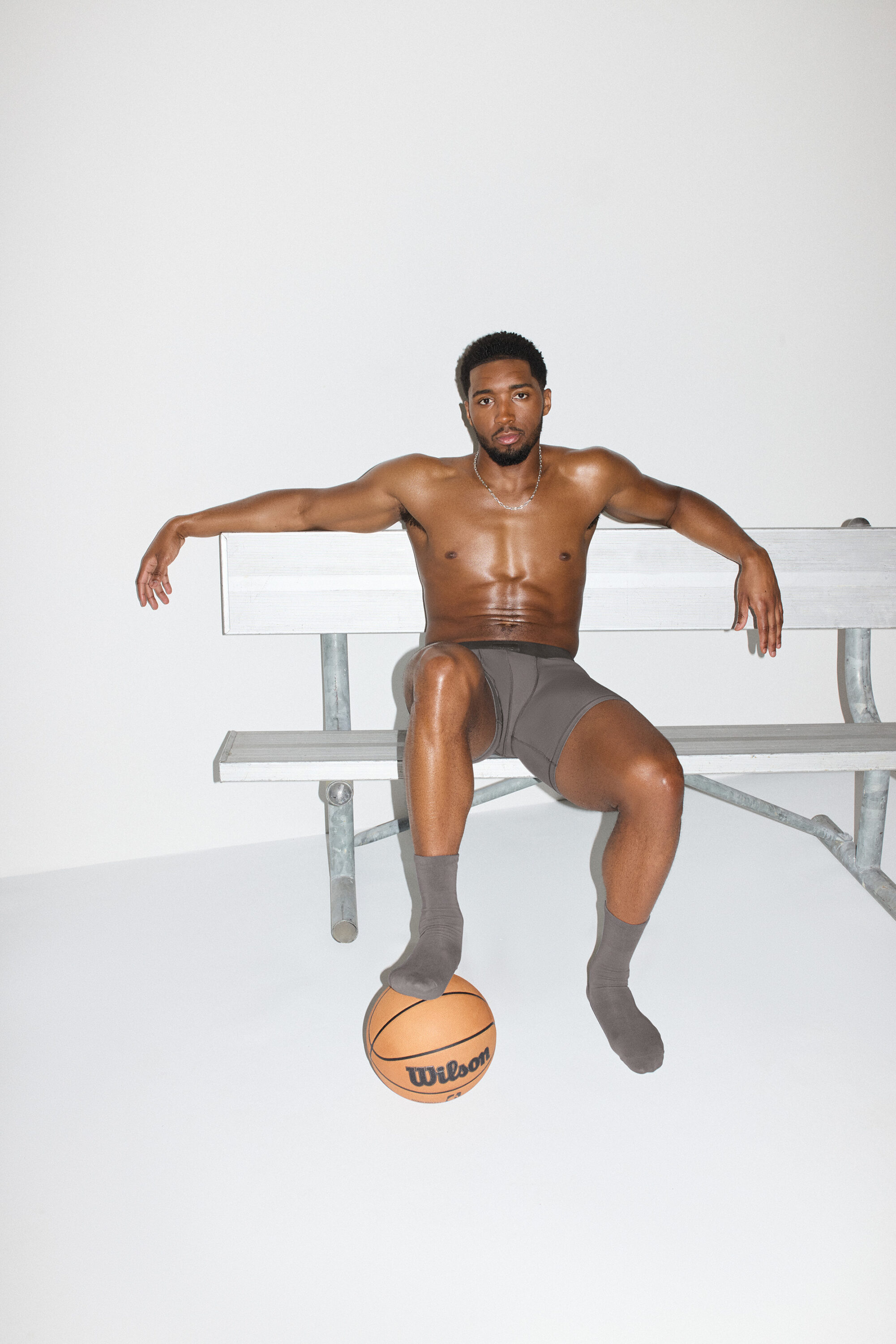 Model Donovan Mitchell seated on a grey bench in grey boxer briefs and socks with one foot on a basketball against a white background.