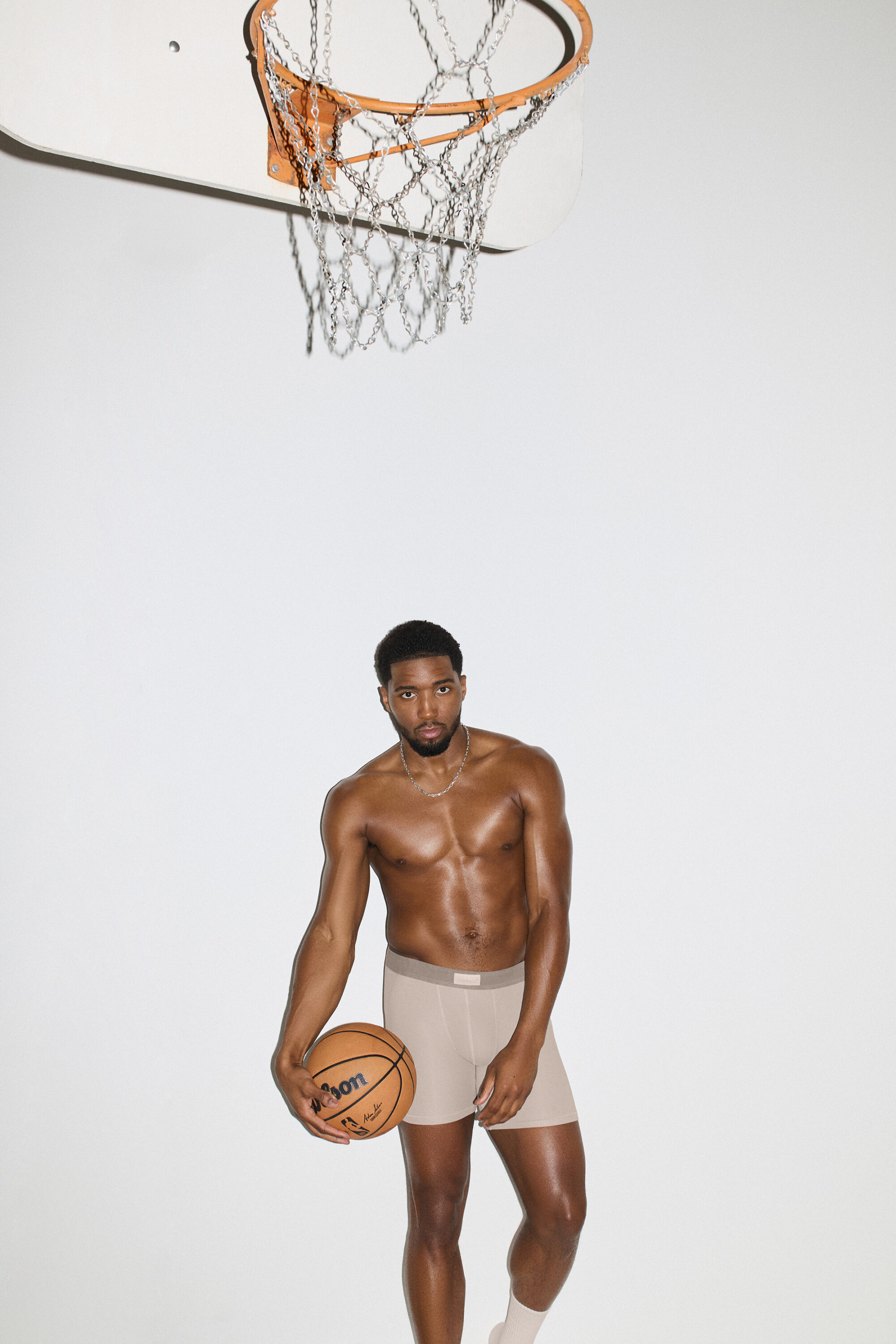 Donovan Mitchell posing shirtless in beige boxer briefs holding a basketball beneath a chain-link hoop against a white seamless background