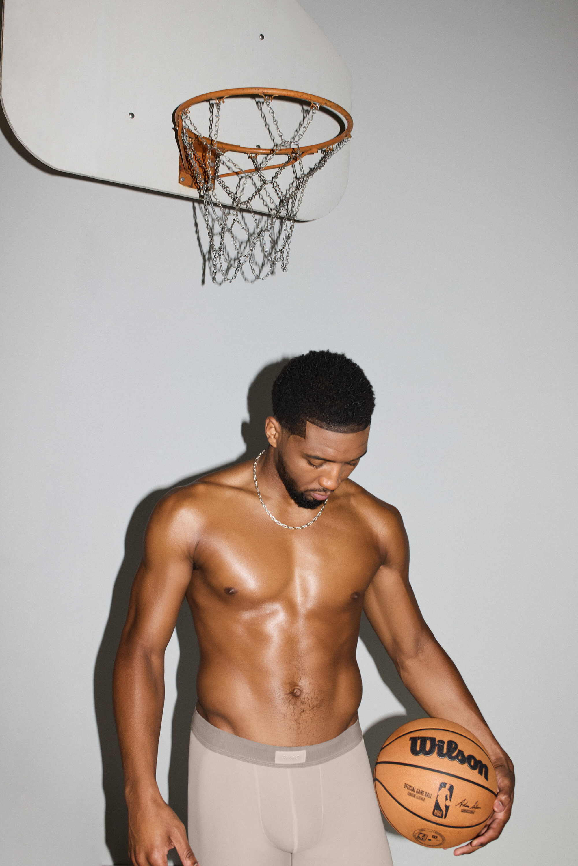 Shirtless black man holding a basketball under a hoop against a plain background for SKIMS.