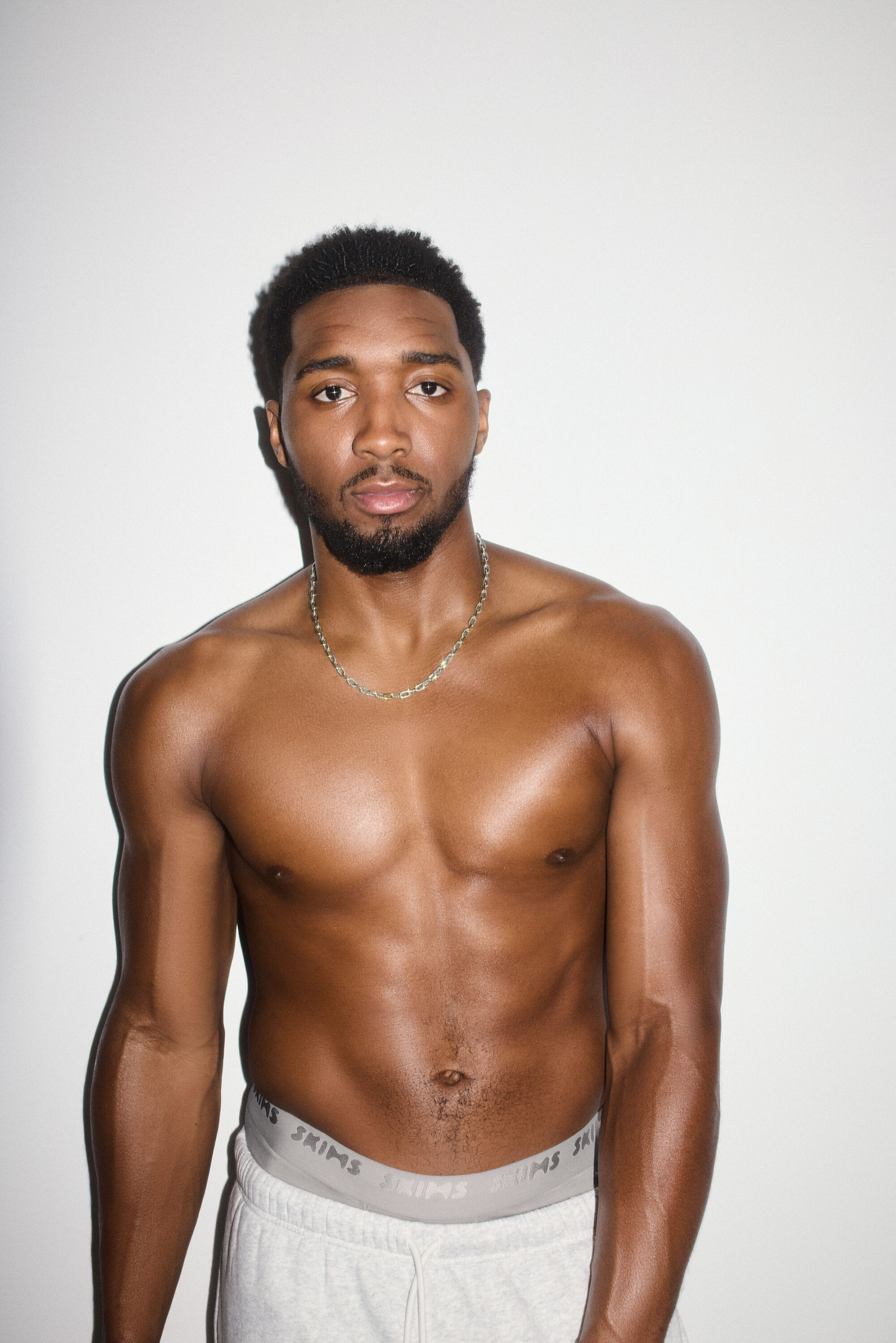 Shirtless man wearing grey sweatpants and silver chain necklace against white background for SKIMS spring 2025 campaign