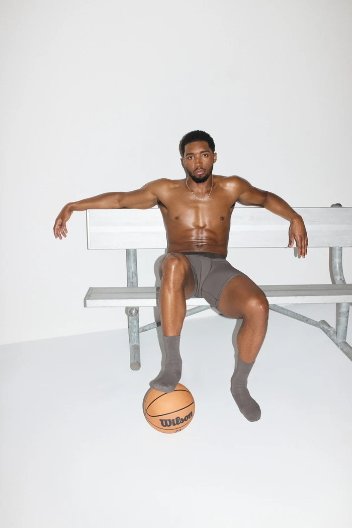 Model Donovan Mitchell seated on a grey bench in grey boxer briefs and socks with one foot on a basketball against a white background.