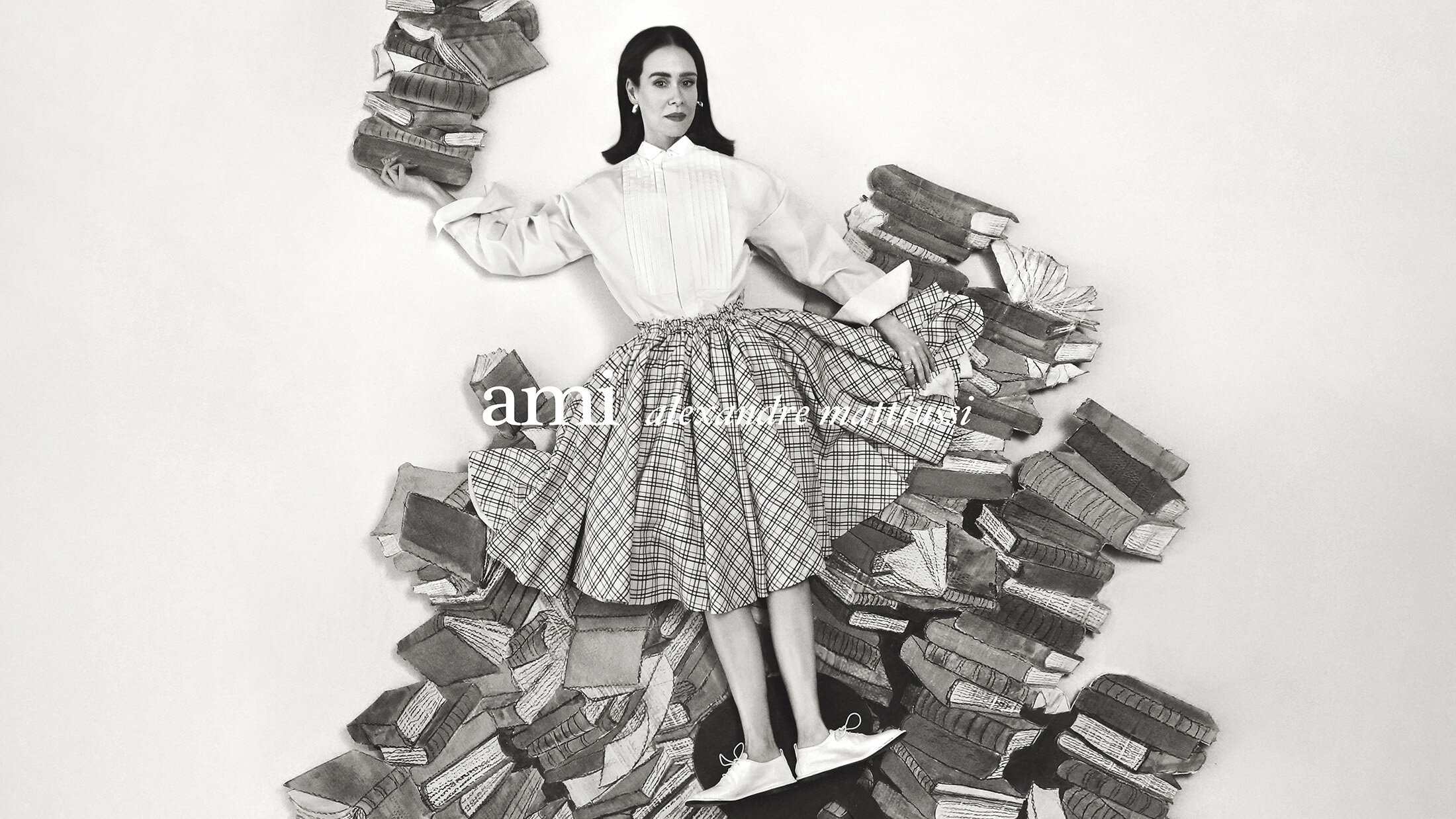 Model Sarah Paulson surrounded by stacks of books wearing a pleated skirt and white shirt for Ami Spring 2025 ad campaign.