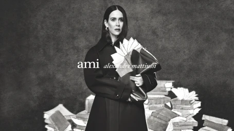 Model Sarah Paulson in a dark belted coat holding a handbag in a black and white fashion campaign image for Ami