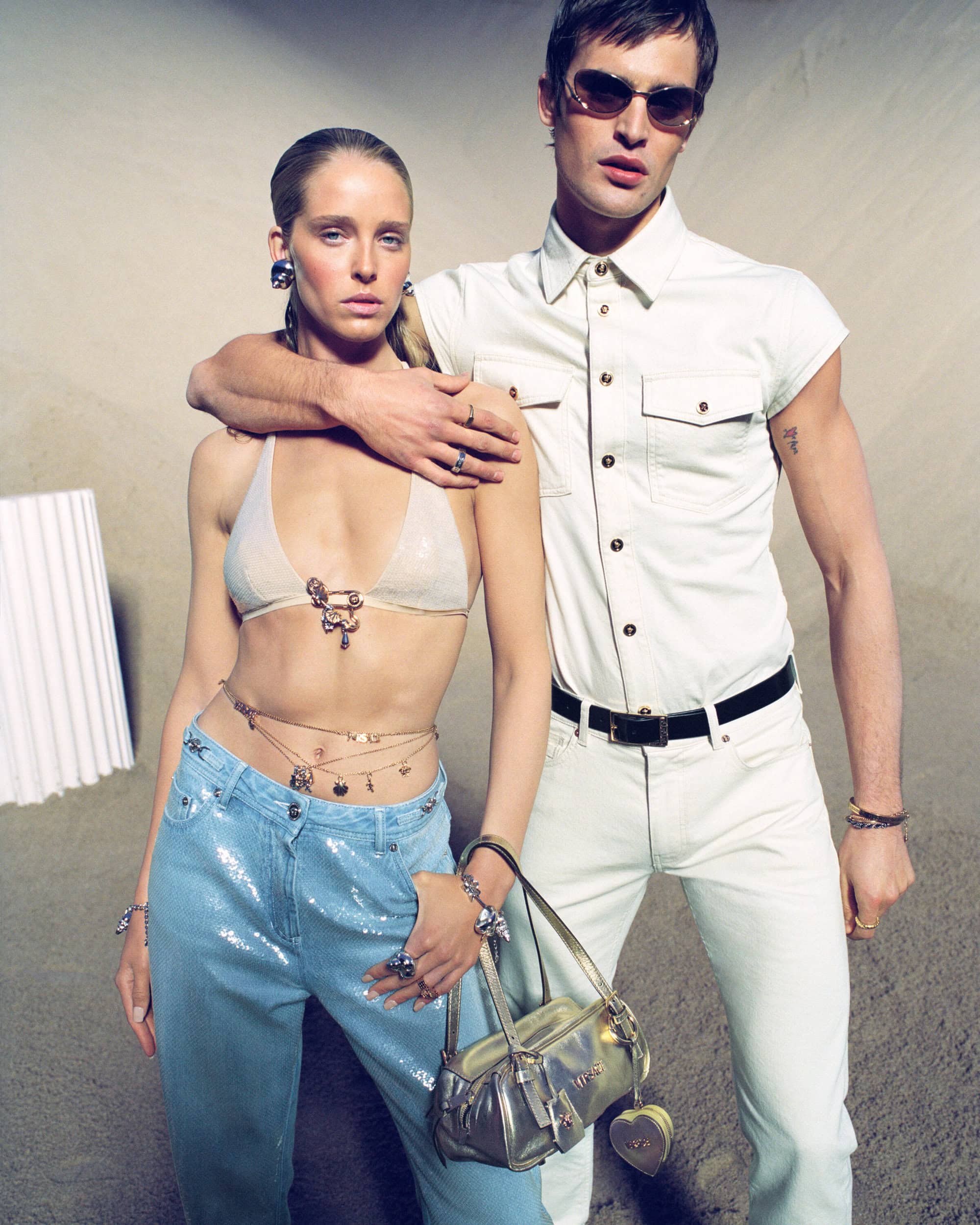 Abby Champion and a male model posing for Versace La Vacanza 2025 ad campaign in a sandy outdoor setting