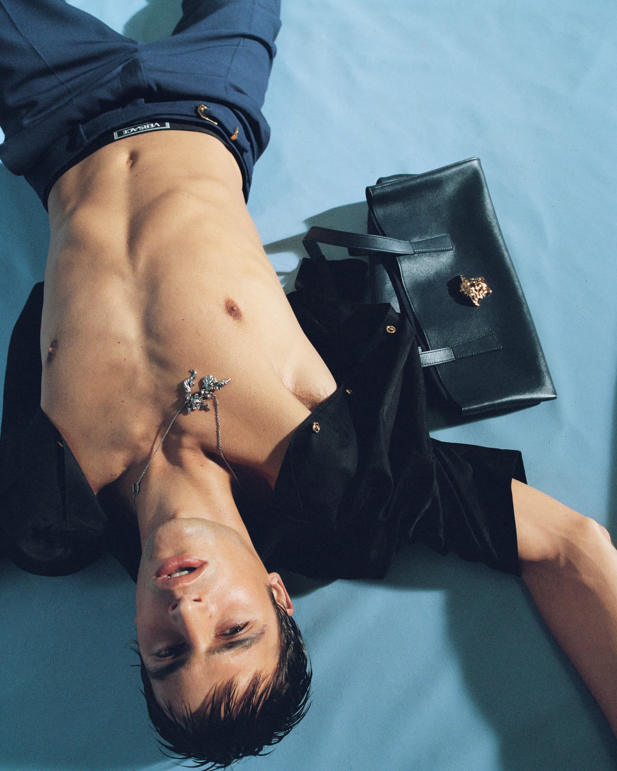 Shirtless male model lying down on a light blue background next to a black handbag, wearing blue trousers.