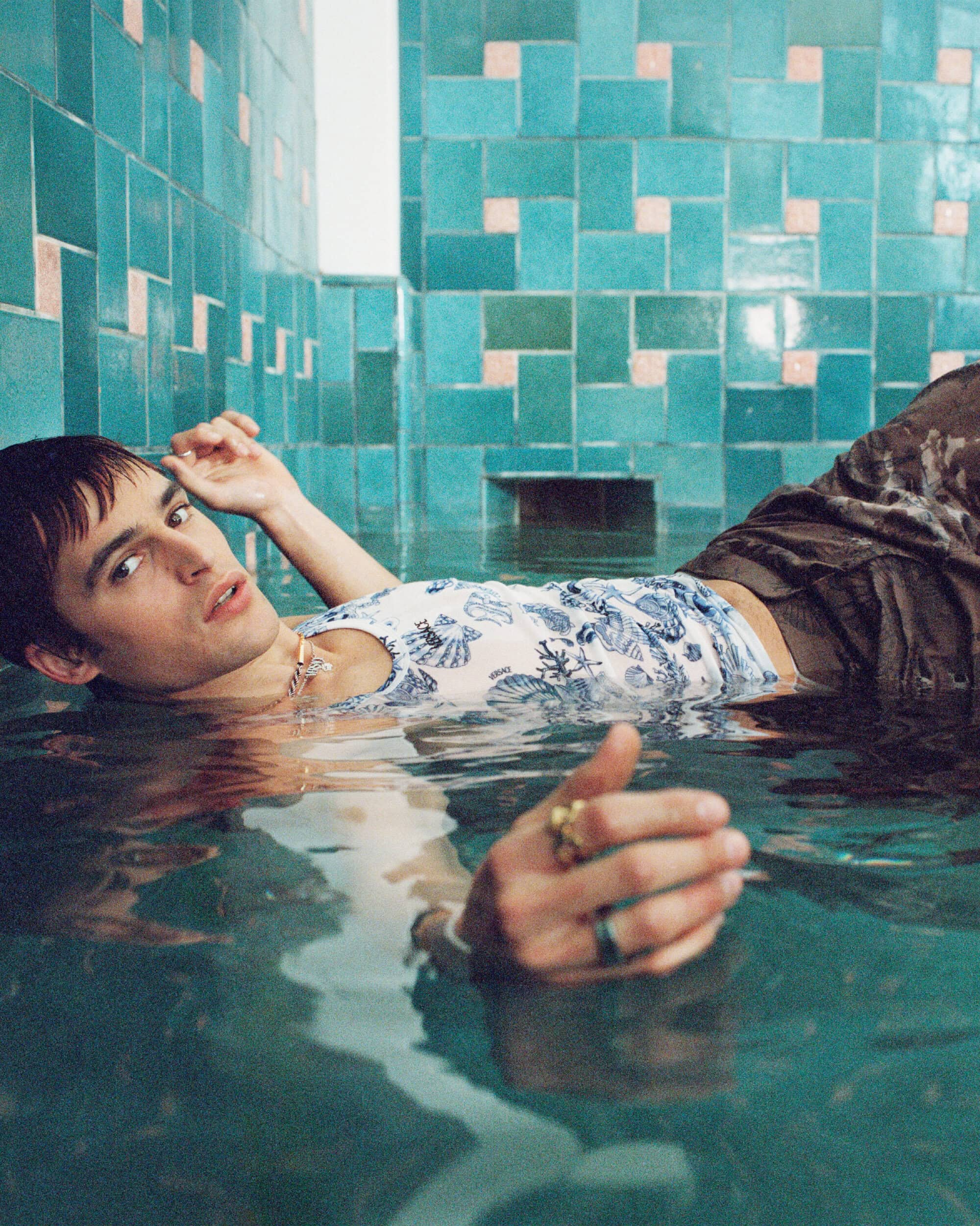 Model floating in a shallow turquoise pool against a tiled wall for Versace La Vacanza 2025 ad campaign