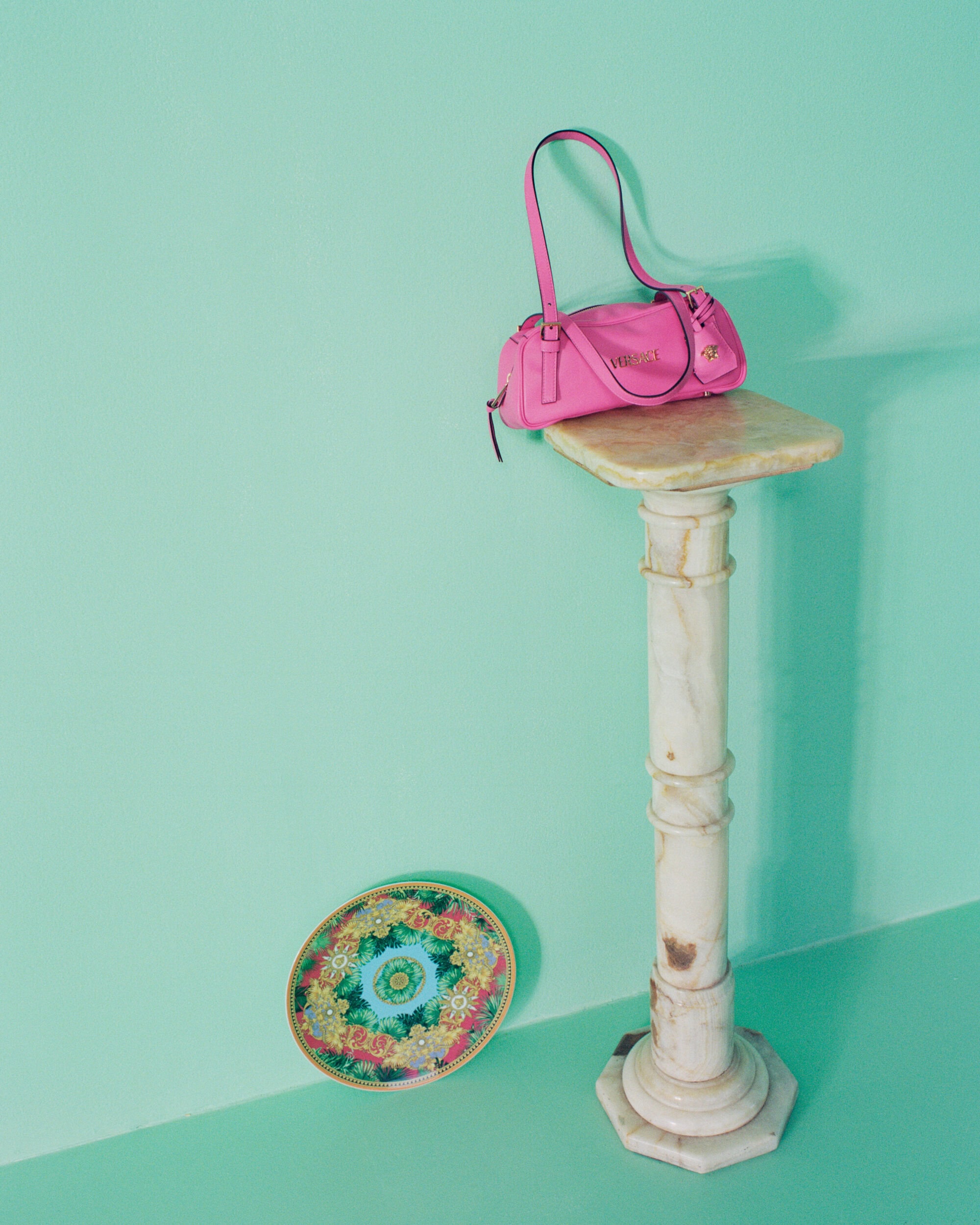 Bright pink leather handbag resting on a marble pedestal against a mint green wall with a decorative patterned plate.