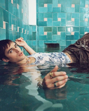 Model floating in a shallow turquoise pool against a tiled wall for Versace La Vacanza 2025 ad campaign