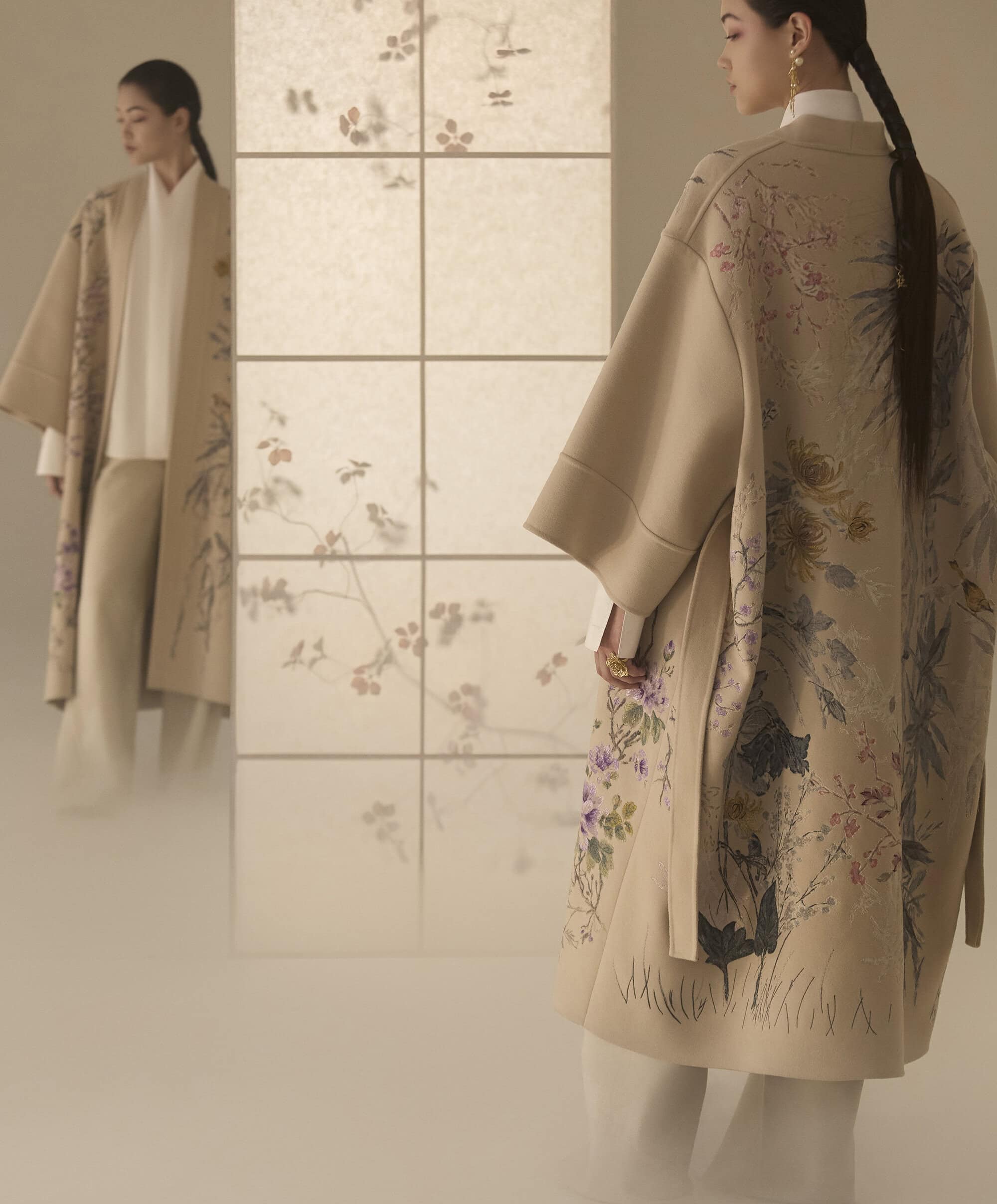 Model wearing a beige embroidered coat standing near an illuminated panel with floral graphics for Dior Fall 2025.
