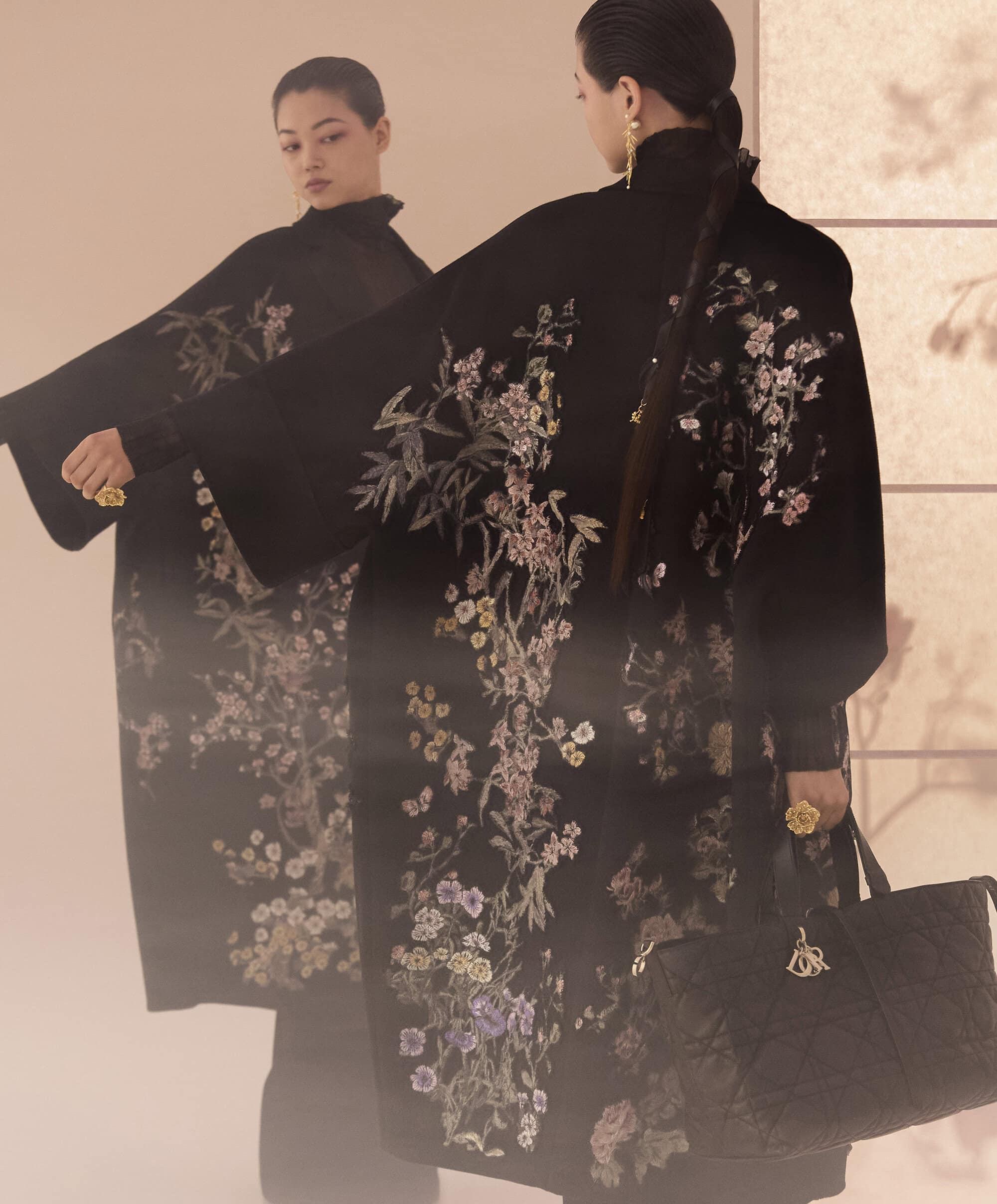 Model in a black embroidered floral coat holding a quilted handbag reflected in a large mirror for the Dior Fall 2025 ad campaign.