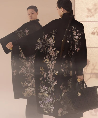 Model in a black embroidered floral coat holding a quilted handbag reflected in a large mirror for the Dior Fall 2025 ad campaign.