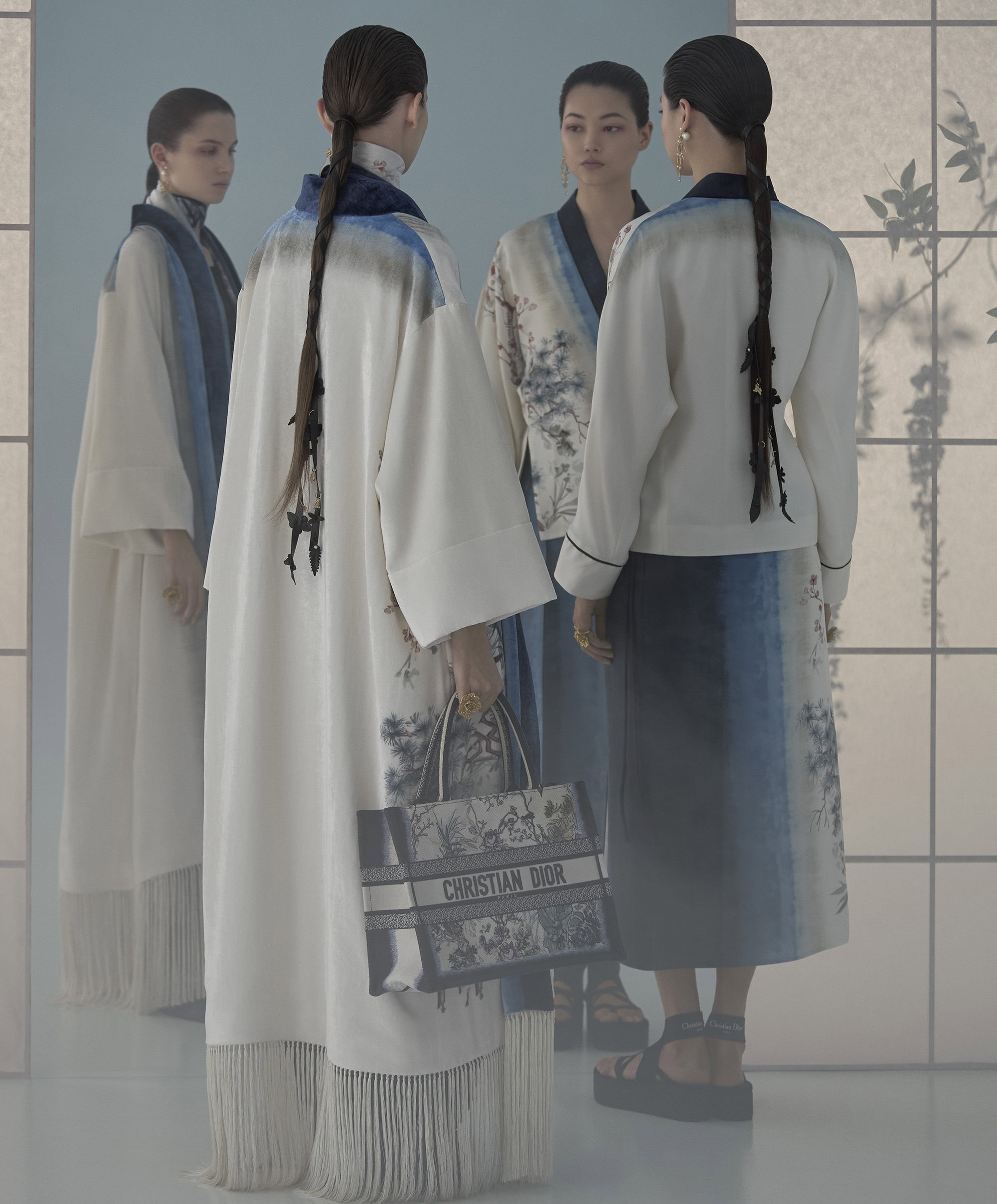 Four female models wearing patterned cream and blue garments holding a Christian Dior tote bag in a studio setting