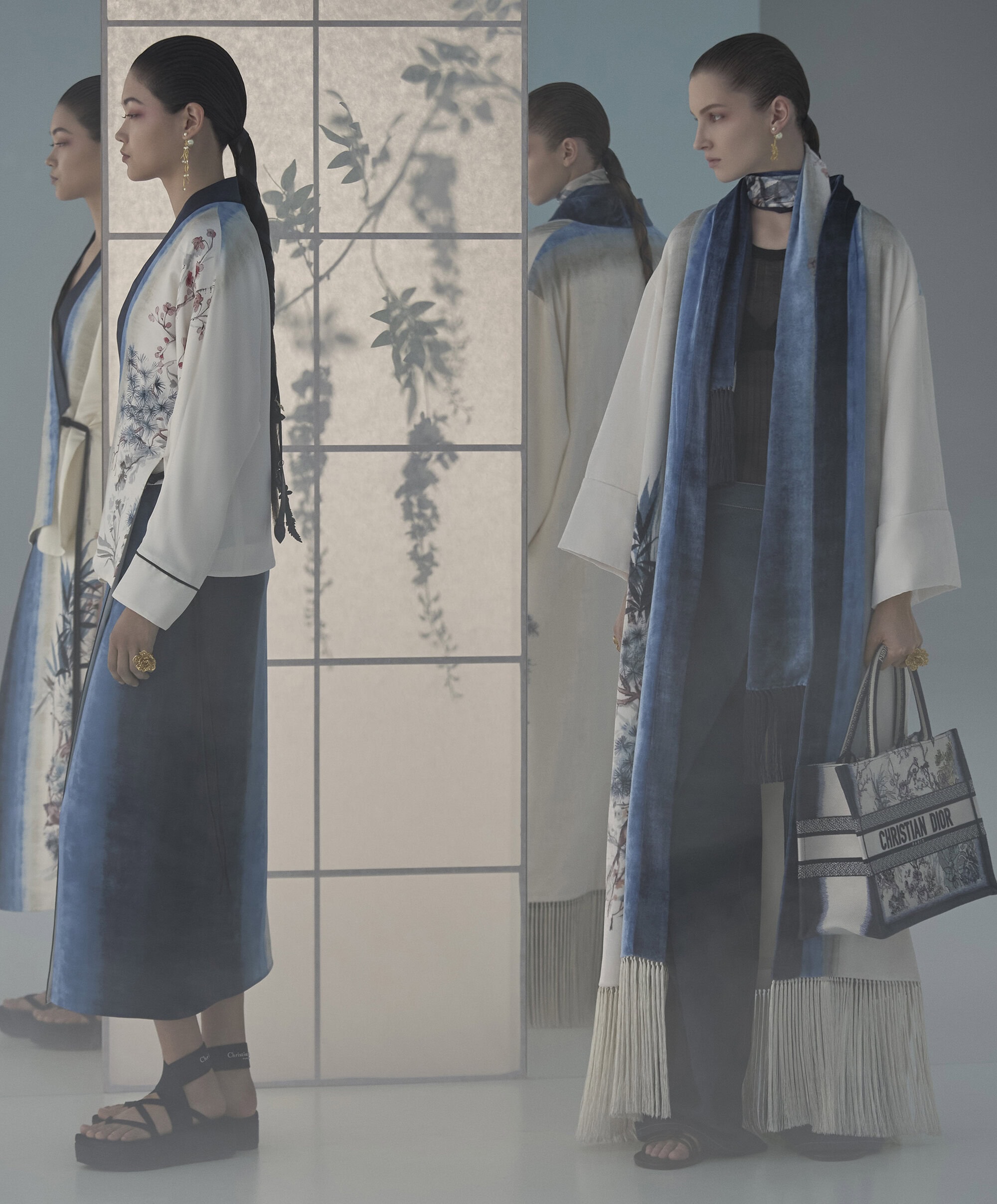 Two female models in flowing coats and dresses standing in a studio with a translucent screen showing botanical shadows