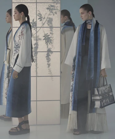 Two female models in flowing coats and dresses standing in a studio with a translucent screen showing botanical shadows