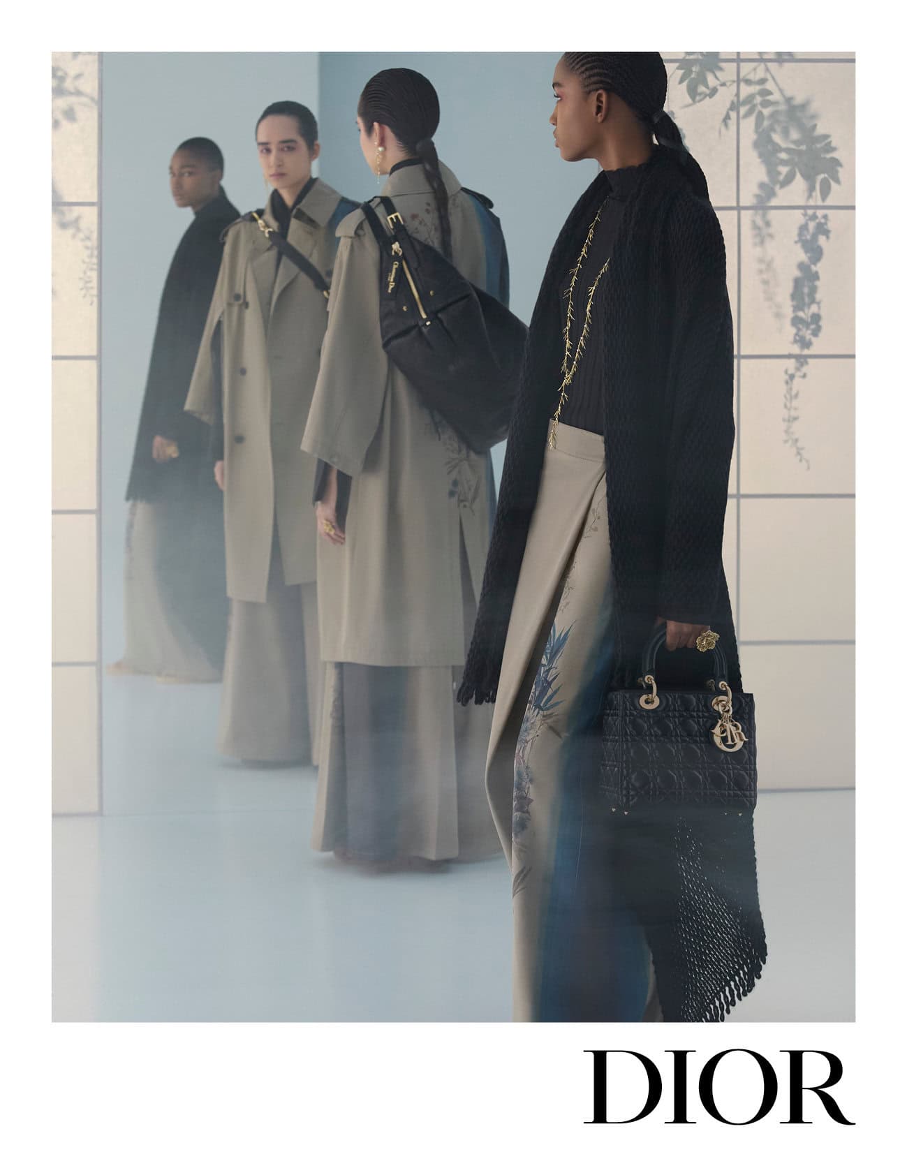 Models in trench coats and black knitwear standing against a pale minimalist studio backdrop for Dior Fall 2025.