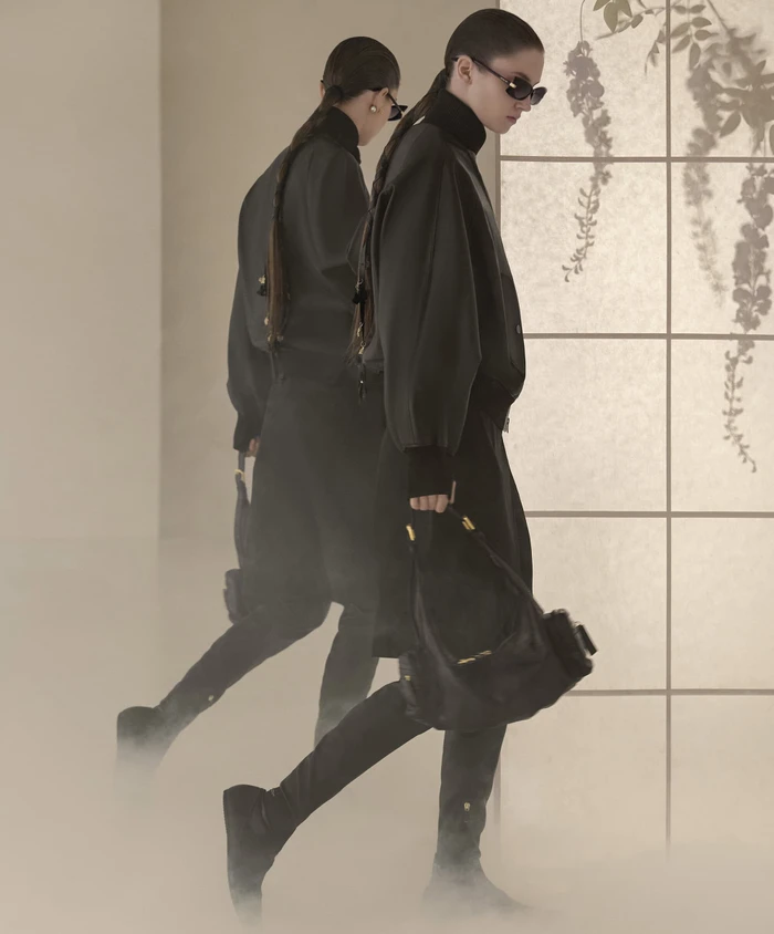Model in dark clothing and sunglasses holding a black handbag walking past a shoji screen