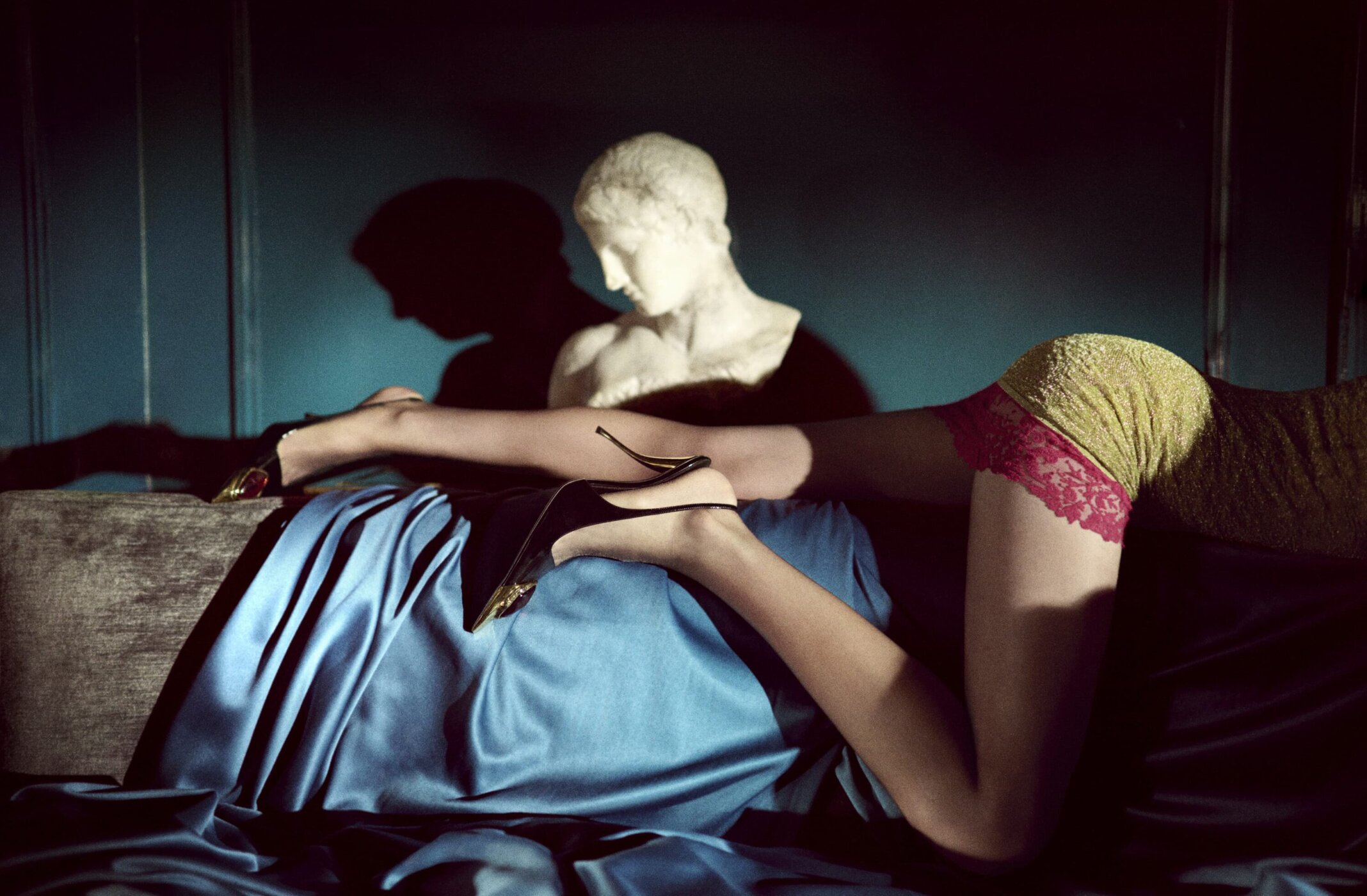 Model draped over a draped blue satin couch with a classical stone bust in the background for Saint Laurent.