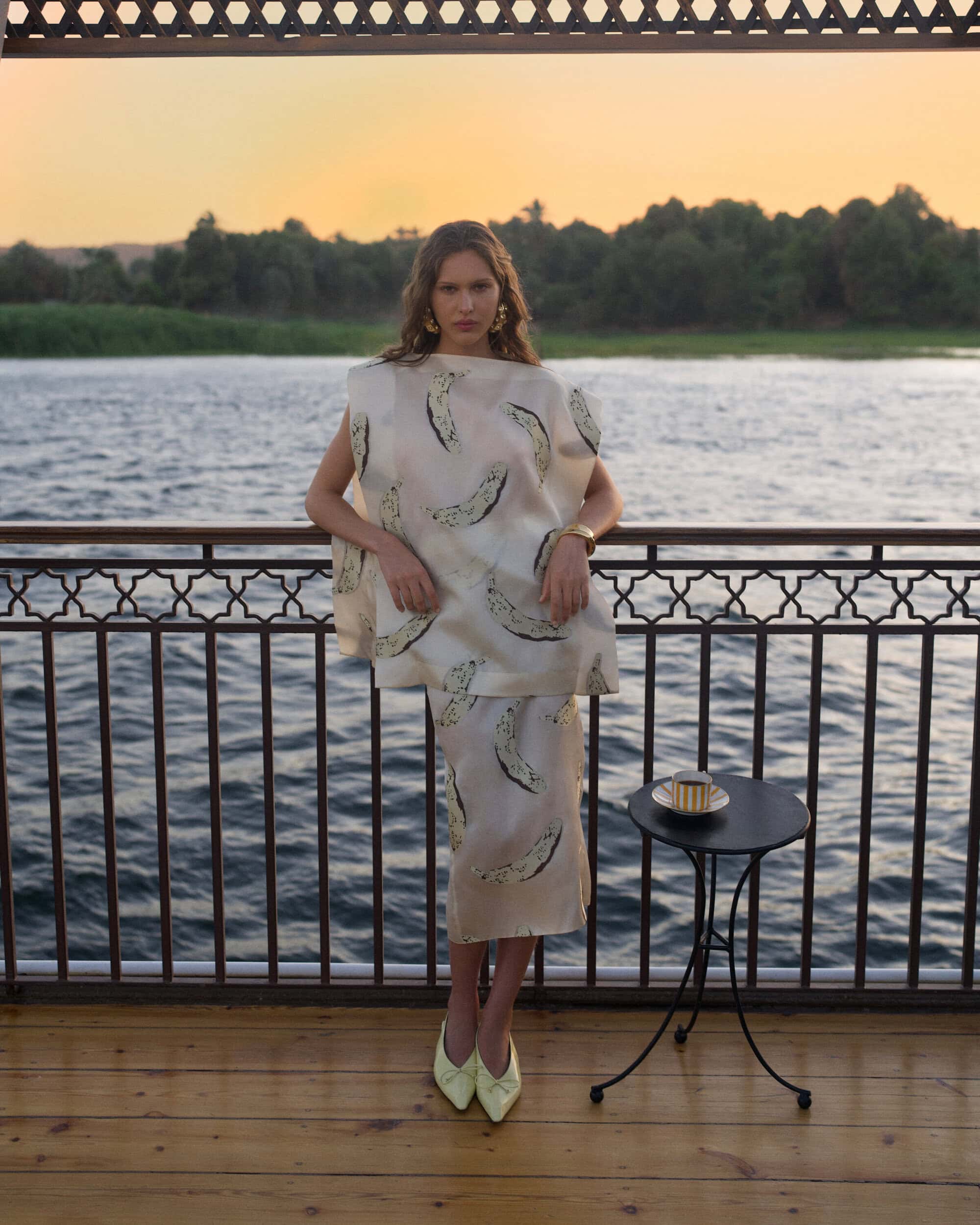 Model leaning on a railing by a river at sunset wearing a printed cream top and skirt with a small table nearby.
