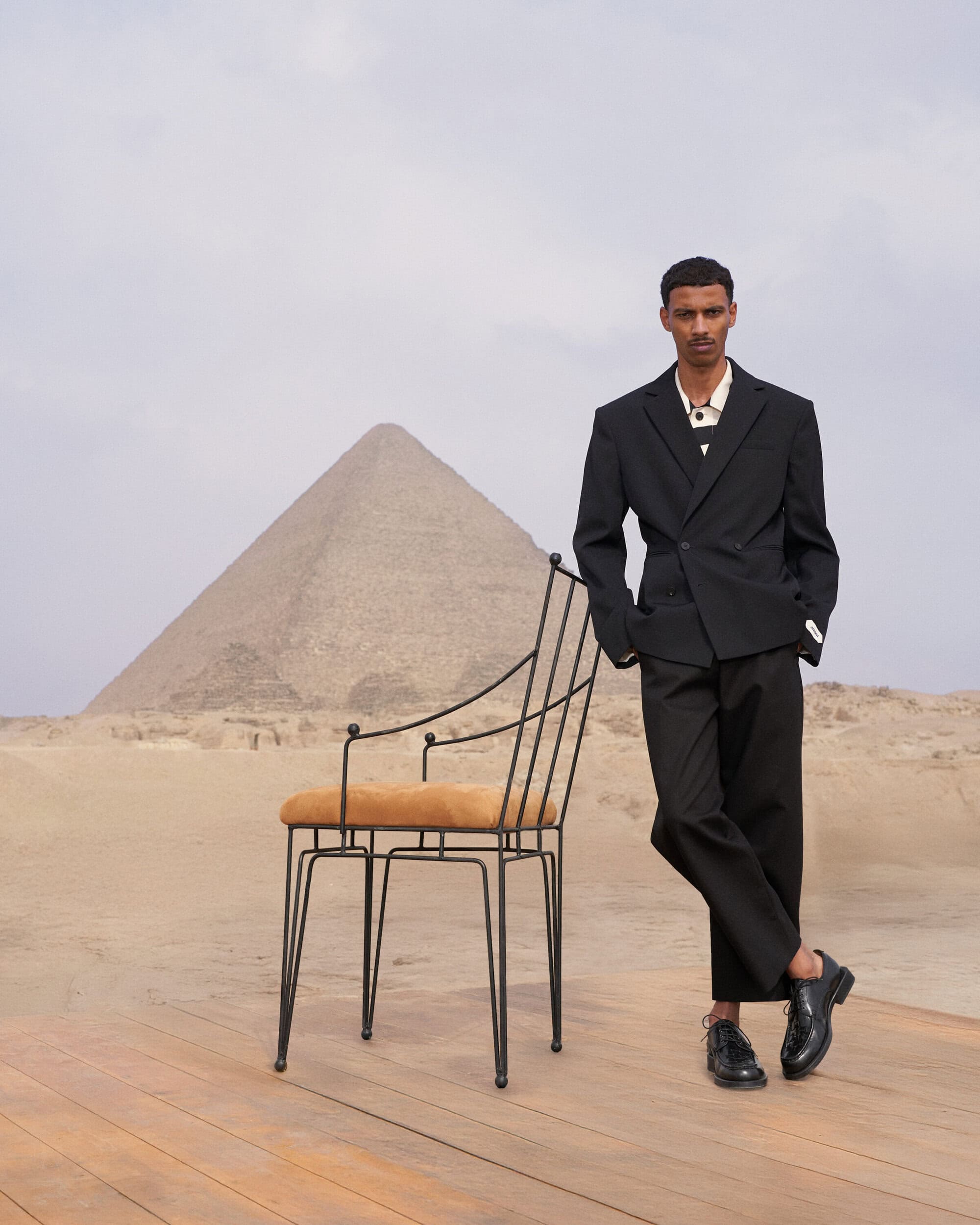 Man wearing a black suit standing next to a wire chair on a wooden platform with a pyramid in the background.