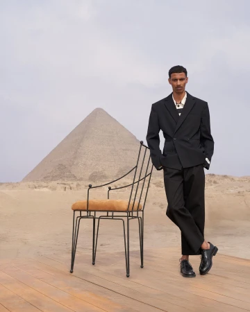 Man wearing a black suit standing next to a wire chair on a wooden platform with a pyramid in the background.