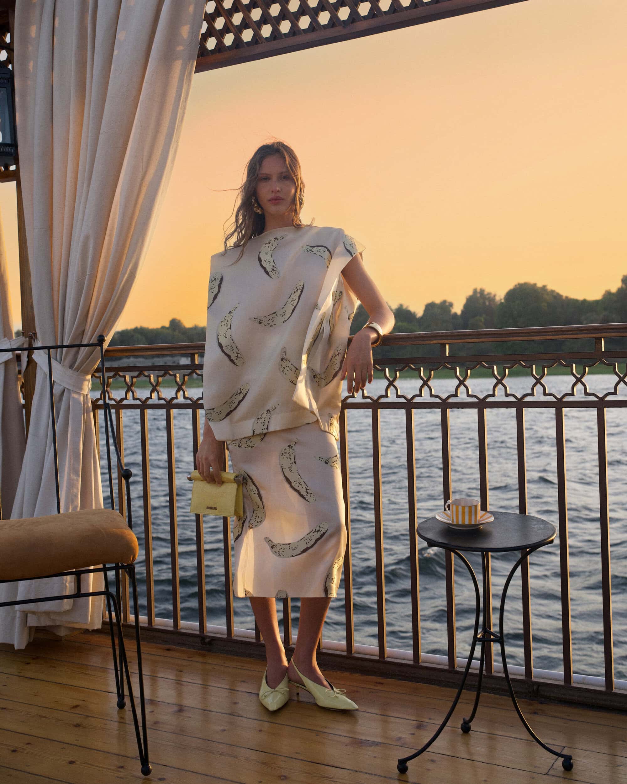 Model standing on a wooden balcony at sunset wearing a patterned top and skirt, holding a small yellow handbag