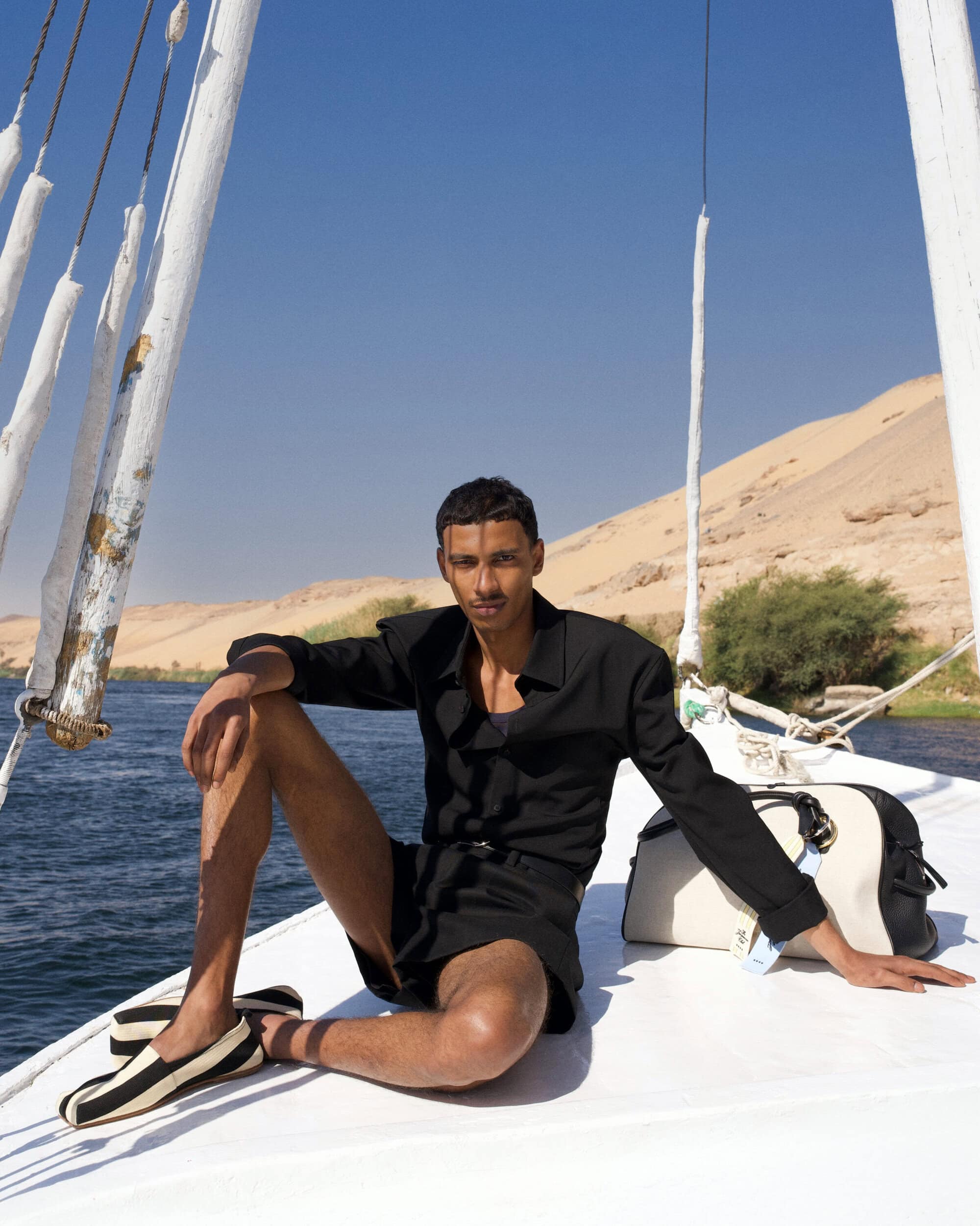 Model sitting on a sailboat deck wearing a black shirt and shorts against water and sandy hills