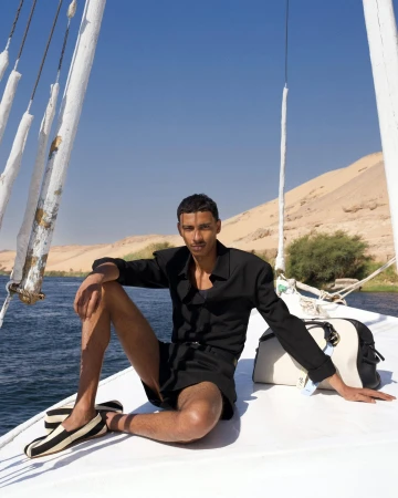 Model sitting on a sailboat deck wearing a black shirt and shorts against water and sandy hills
