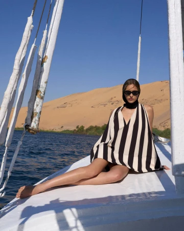 Model wearing striped dress and headscarf seated on white boat deck with blue water and sandy shore behind