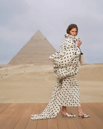 A female model wearing a polka dot quilted coat and matching shoes walks on a wooden platform with a large pyramid in the desert background.