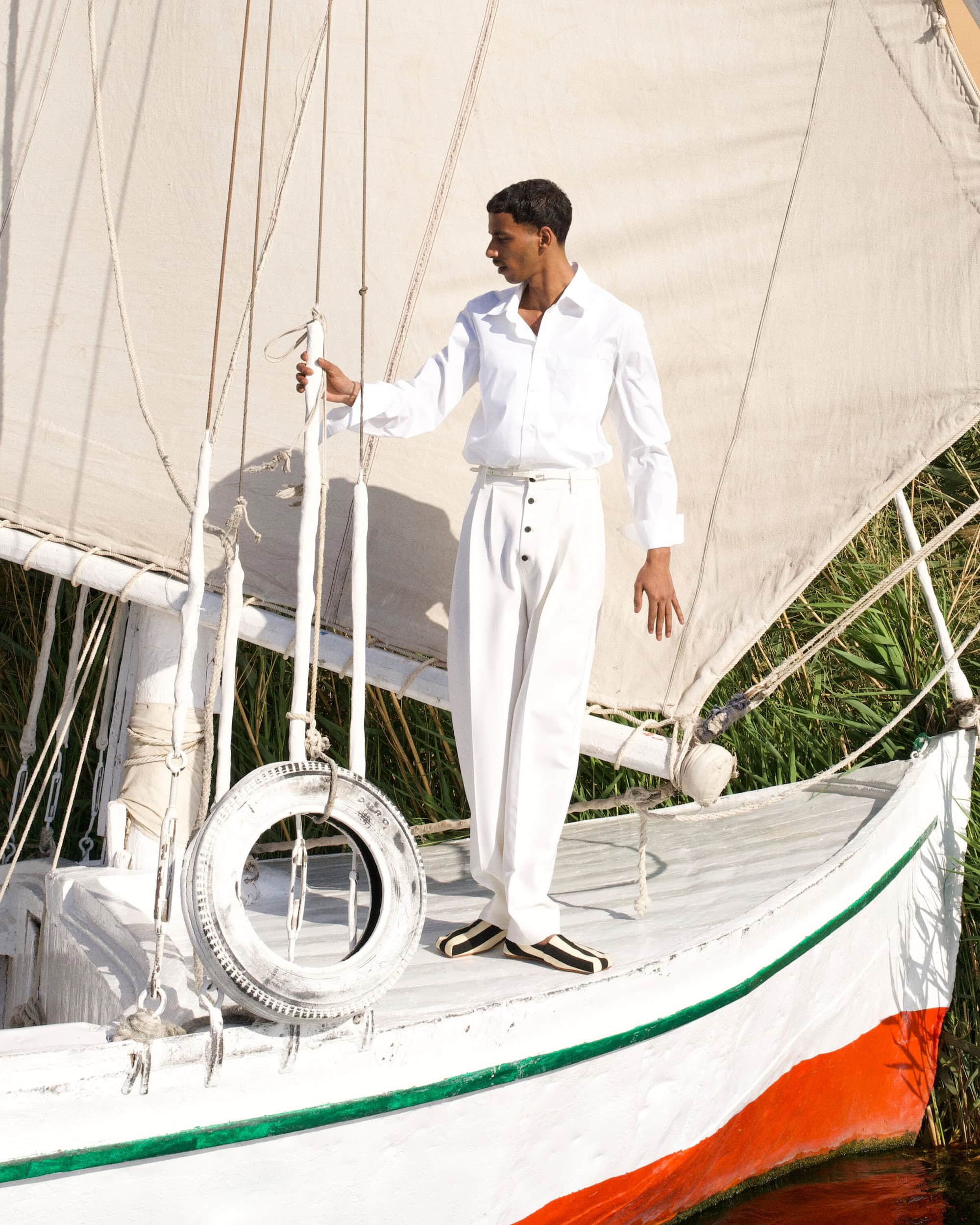 Man wearing a white shirt and trousers standing on the deck of a sailboat with a white sail