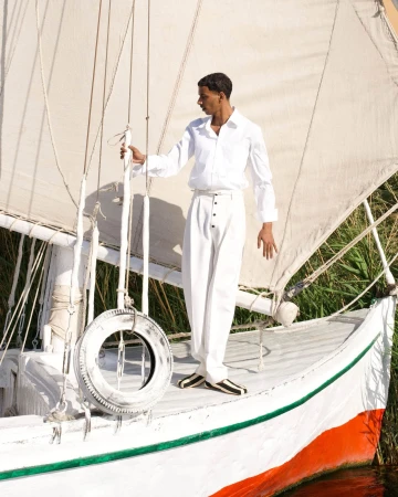 Man wearing a white shirt and trousers standing on the deck of a sailboat with a white sail