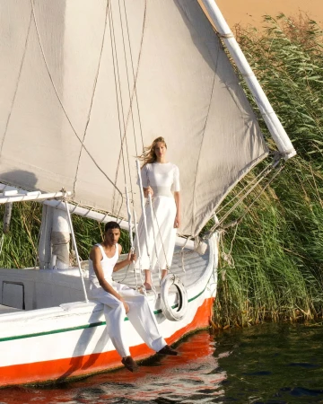Two models in white clothing on a traditional wooden sailboat among tall reeds for Jacquemus La Croisière campaign.