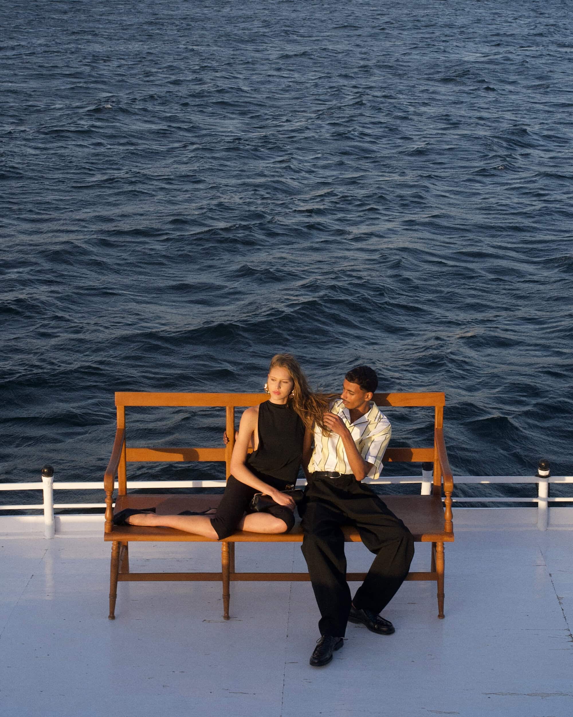A man and woman seated together on a wooden bench outdoors against a backdrop of deep blue water.