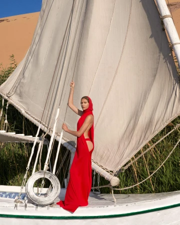 Model in a red dress standing on a traditional boat with a large white sail against a sandy background