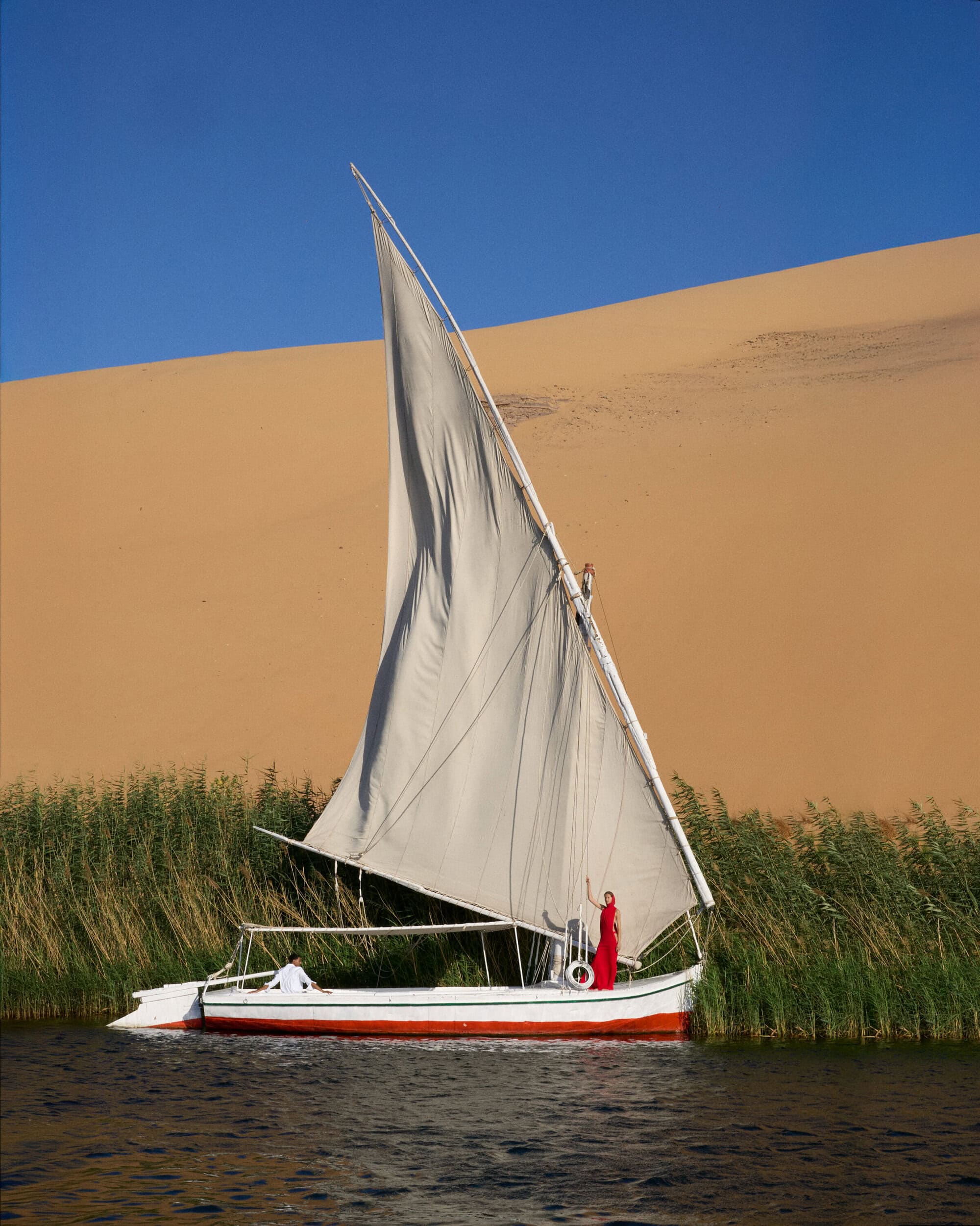 Traditional wooden sailboat with a large white triangular sail on calm water next to a tall sand dune and green reeds