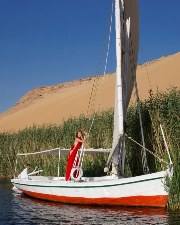 Model in a red dress standing on a white sailboat docked among tall river reeds with a sandy desert dune and clear blue sky behind.