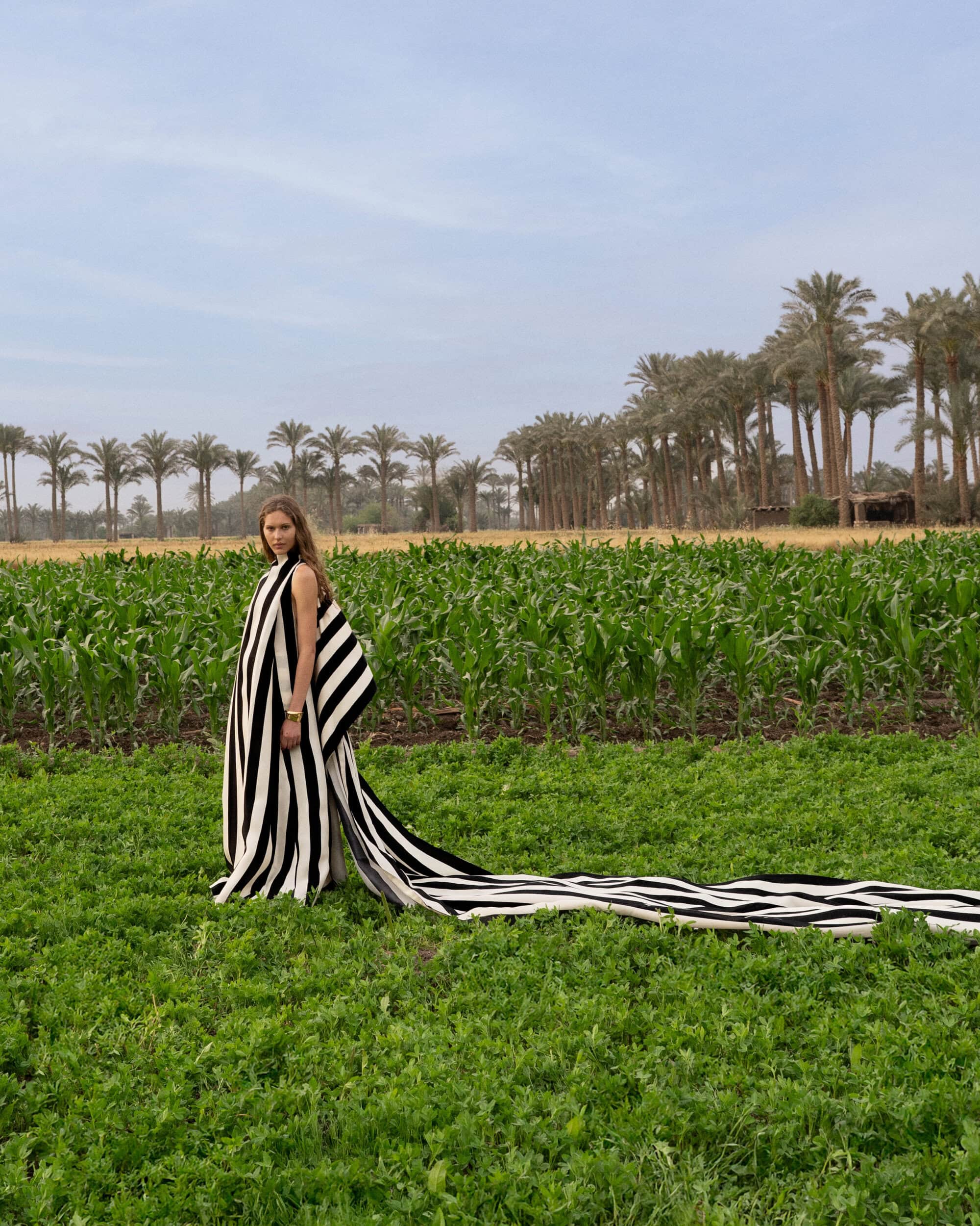 Model in a black and white striped gown with a long trailing cape standing in a lush green agricultural field with palm trees.