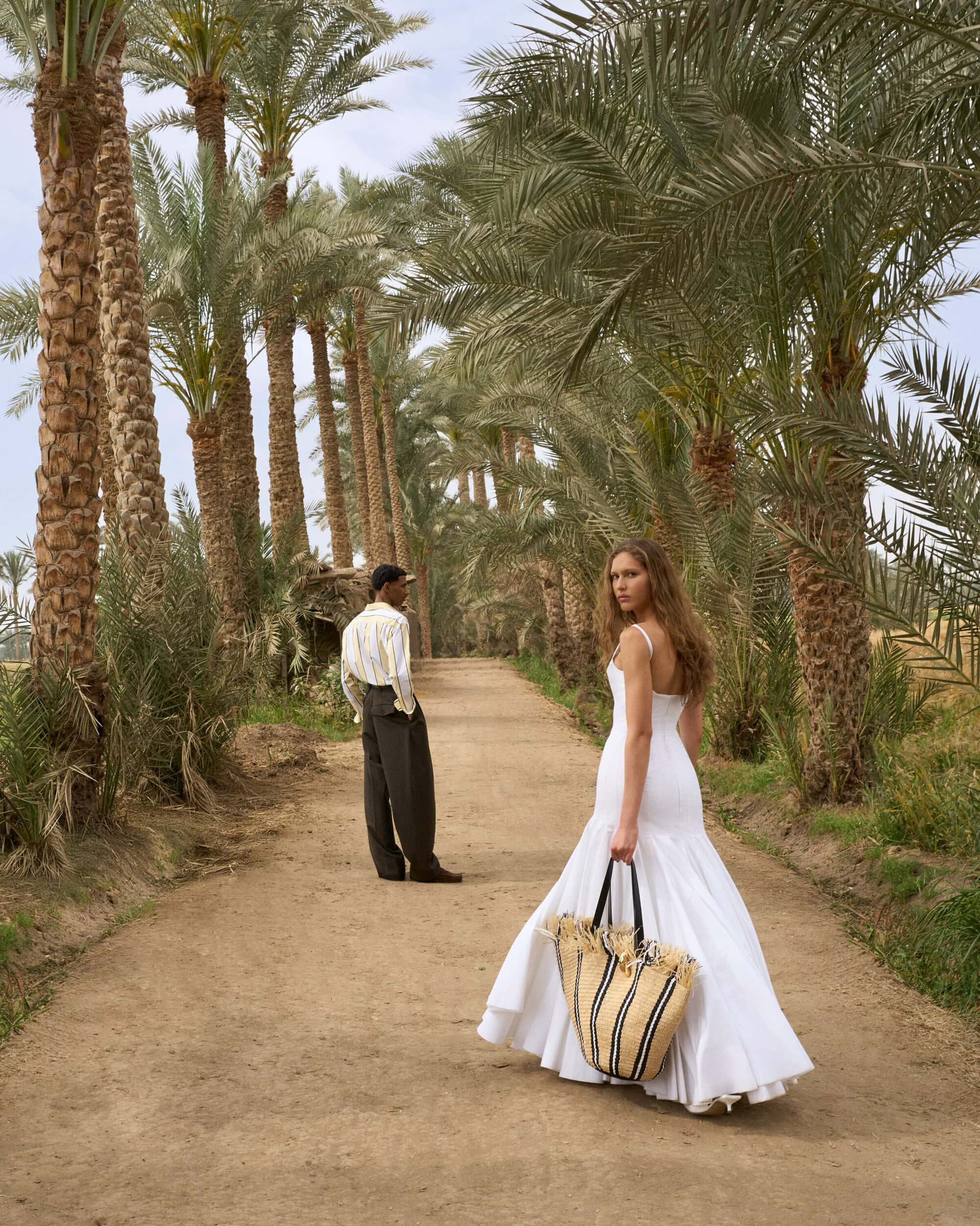 Woman in a long white dress holding a straw tote bag walking down a palm tree-lined dirt path with a man standing in the background.