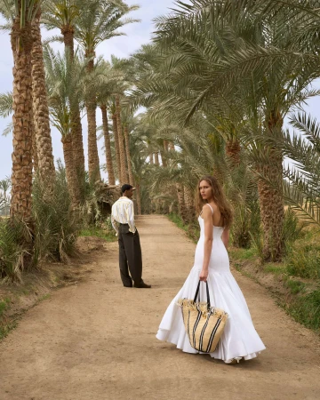Woman in a long white dress holding a straw tote bag walking down a palm tree-lined dirt path with a man standing in the background.
