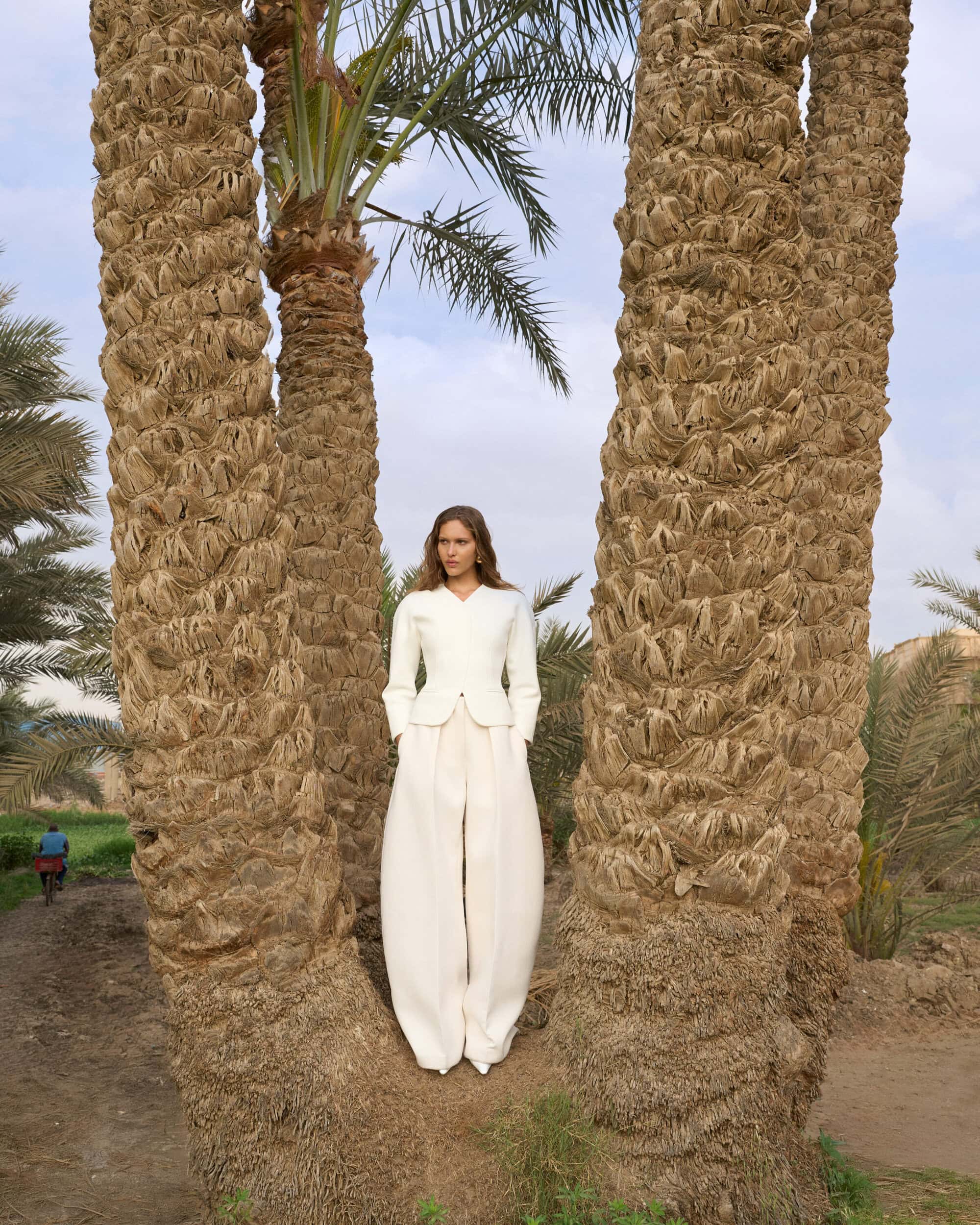 Model standing between tall palm tree trunks wearing a white structured top and wide trousers outdoors