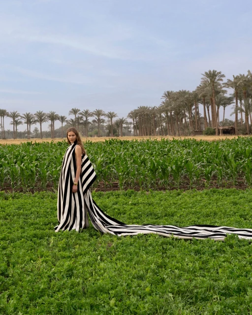 Model in a black and white striped gown with a long trailing cape standing in a lush green agricultural field with palm trees.