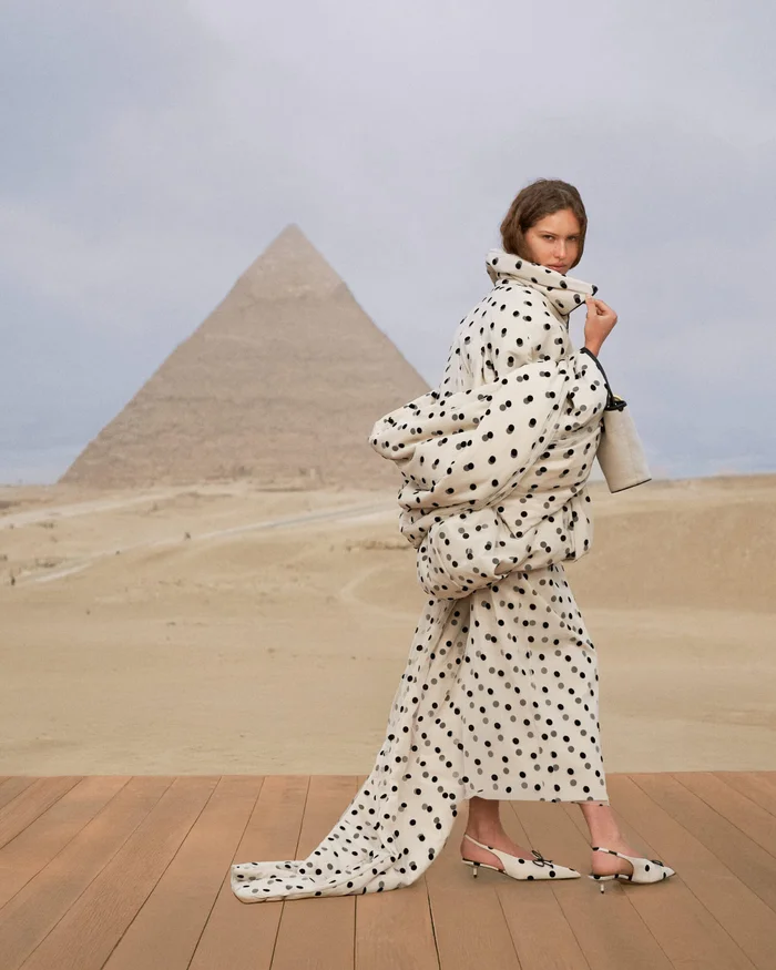 A female model wearing a polka dot quilted coat and matching shoes walks on a wooden platform with a large pyramid in the desert background.