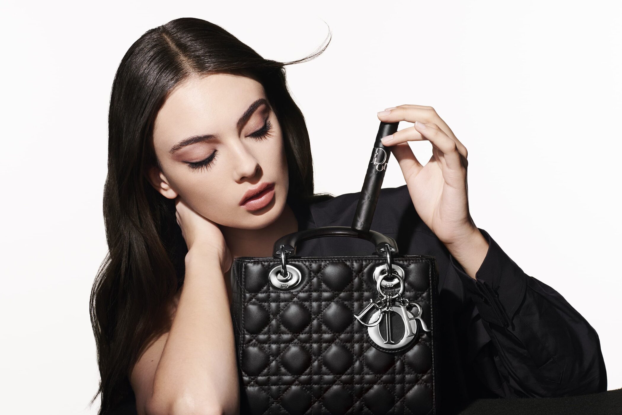A dark-haired woman holding a black quilted handbag against a stark white background for Dior Beauty 2025 ad campaign