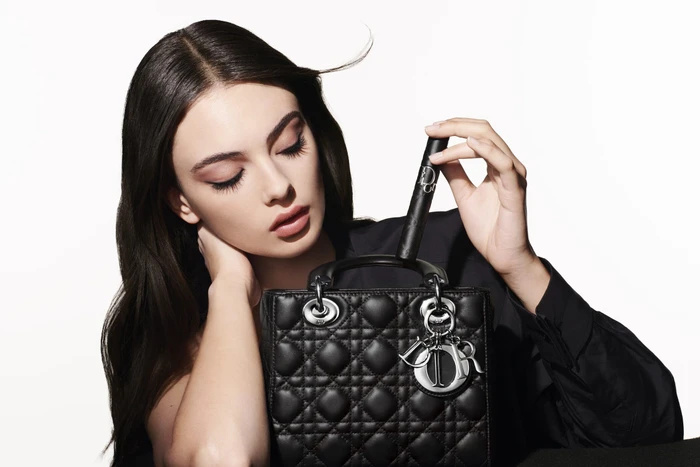A dark-haired woman holding a black quilted handbag against a stark white background for Dior Beauty 2025 ad campaign