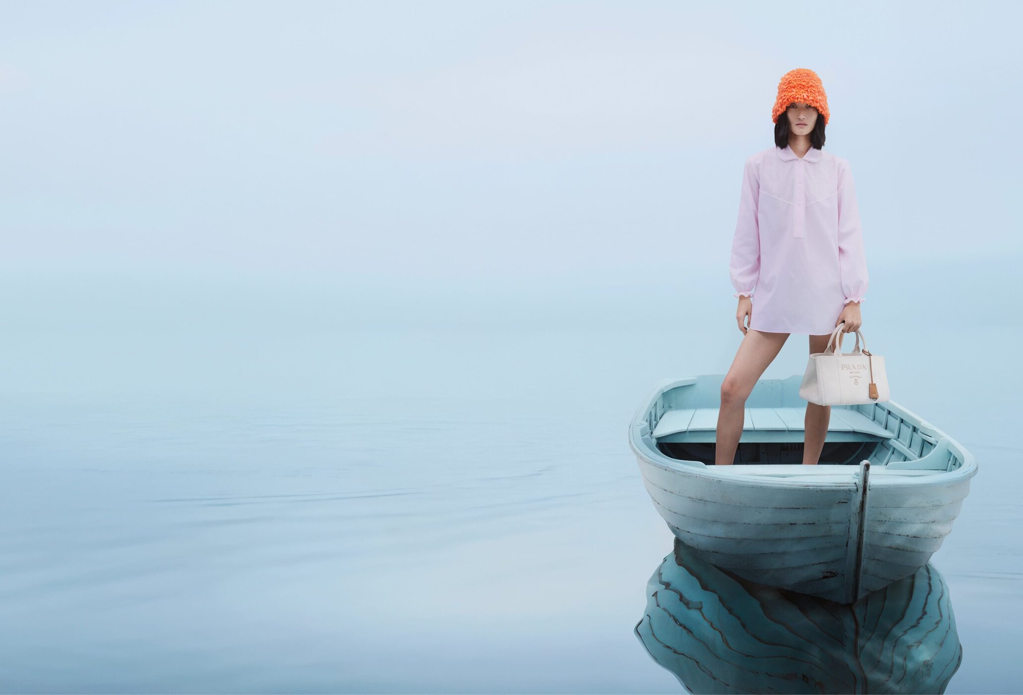 Model wearing an orange fuzzy hat and pink shirt standing in a light blue boat on a calm foggy lake holding a white handbag