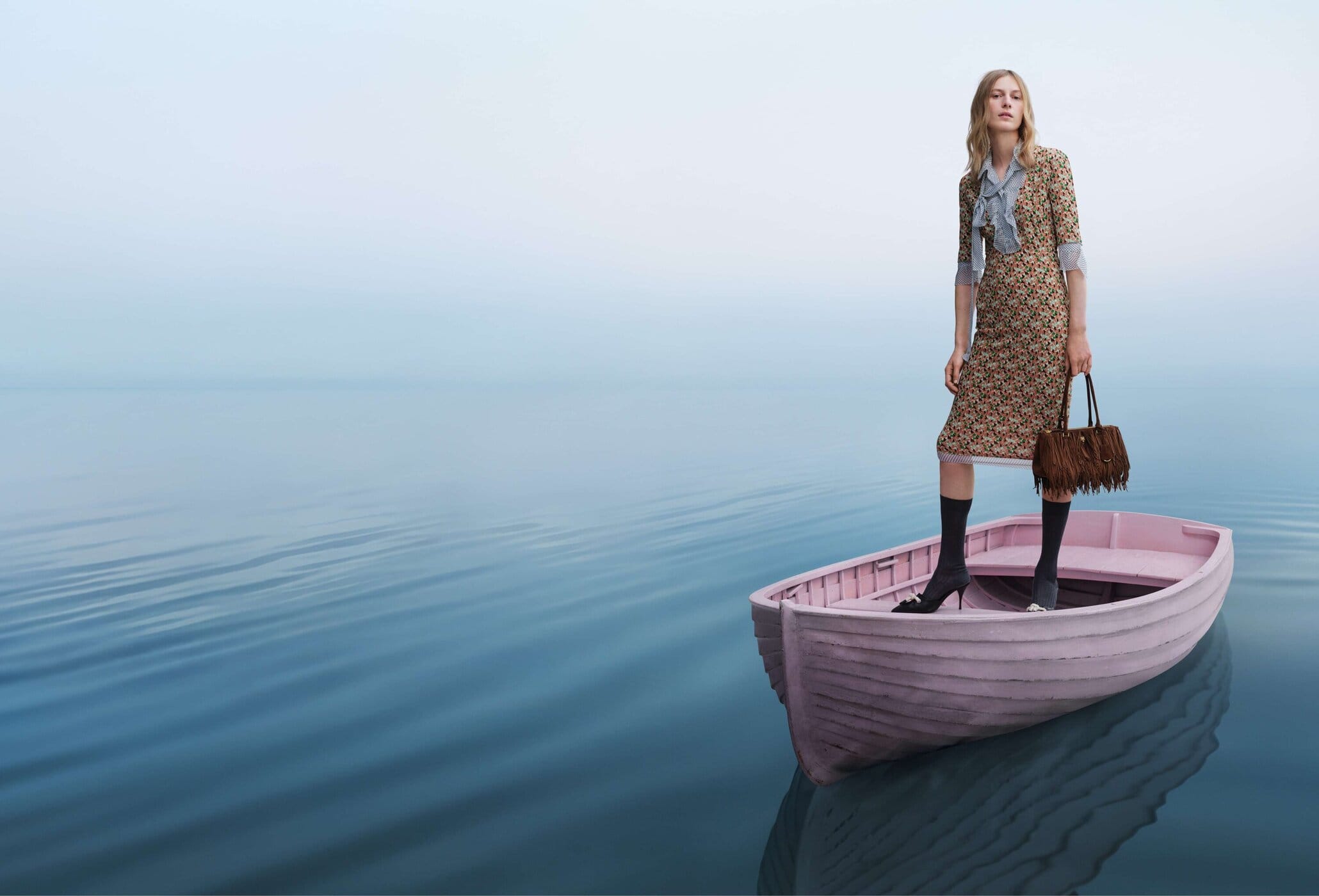 Model standing inside a pink wooden rowboat on calm water holding a brown fringed handbag