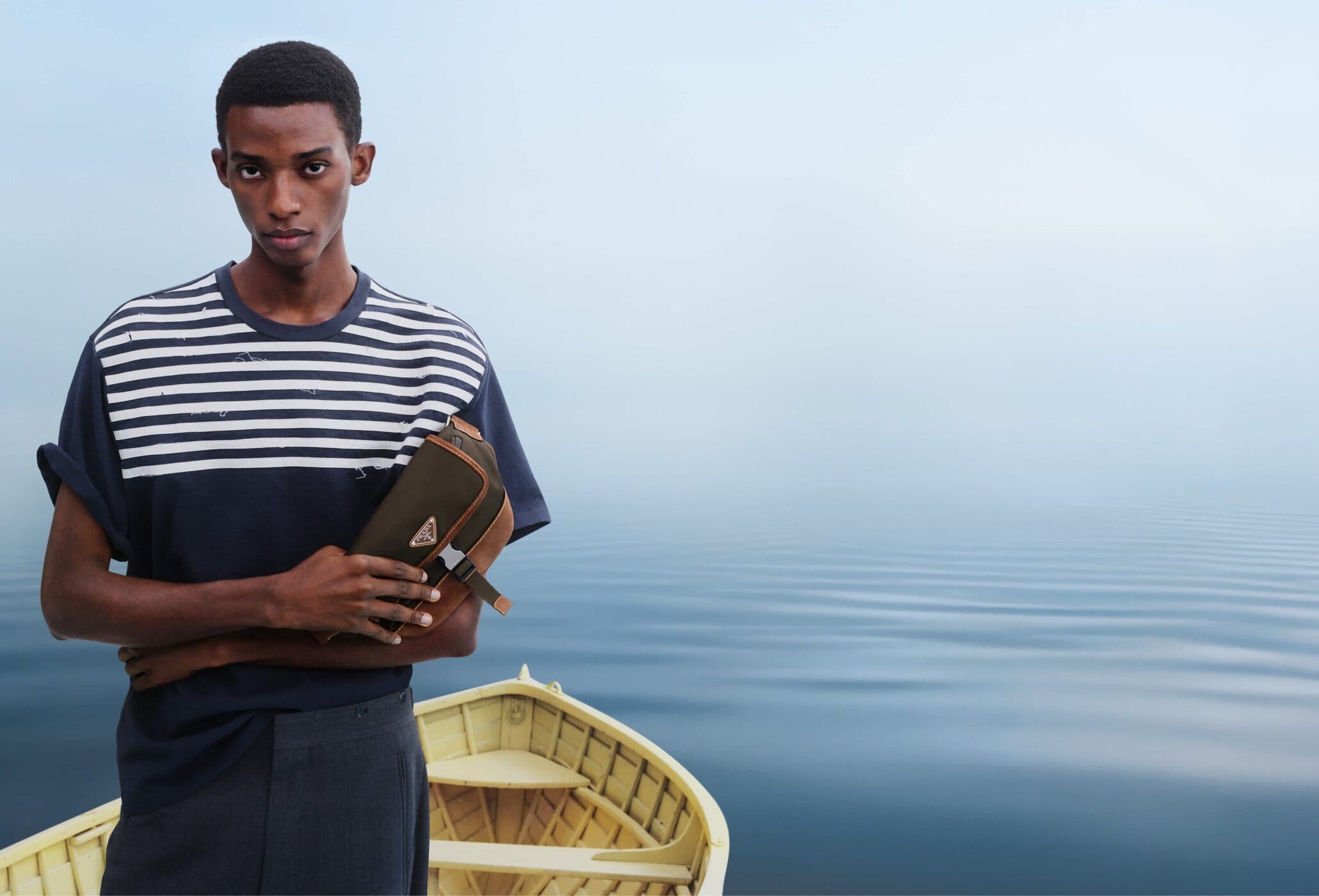 A Black man standing in a wooden boat on a misty body of water, wearing a striped t-shirt and holding an olive green crossbody bag.