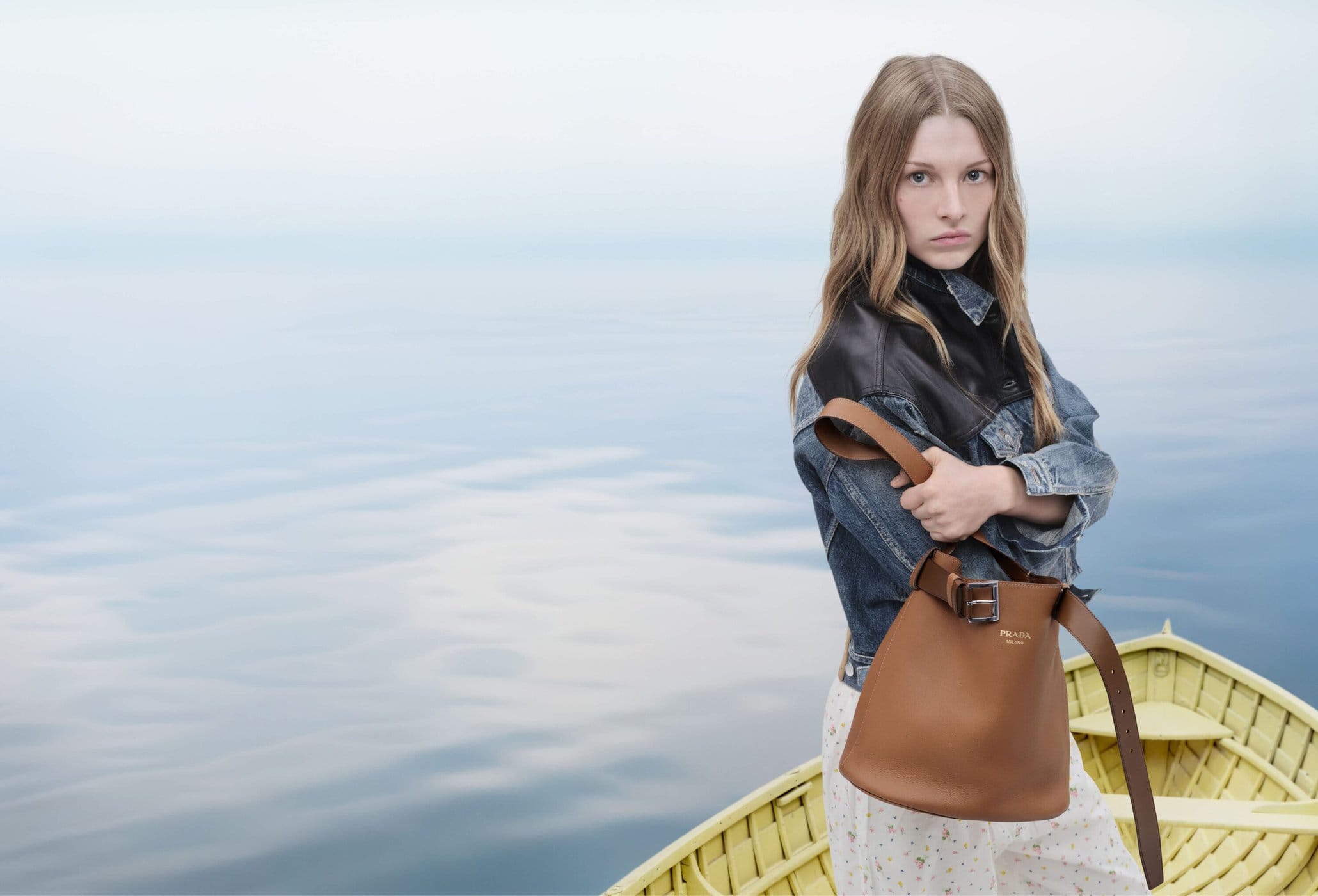 Model wearing a denim and leather jacket holding a brown leather bucket bag while standing in a yellow wooden boat on open water